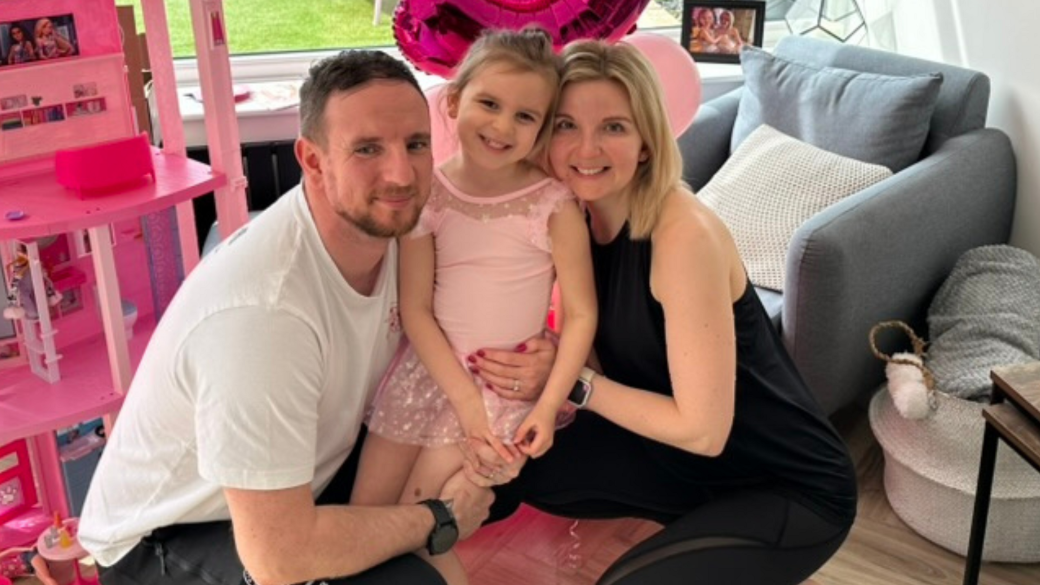 Tor, with her husband, Joe and daughter Isla Rose, both parents are crouching down, holding their daughter, wearing a pink dress, in a room, with pink toys and furniture. They are all looking at the camera. 