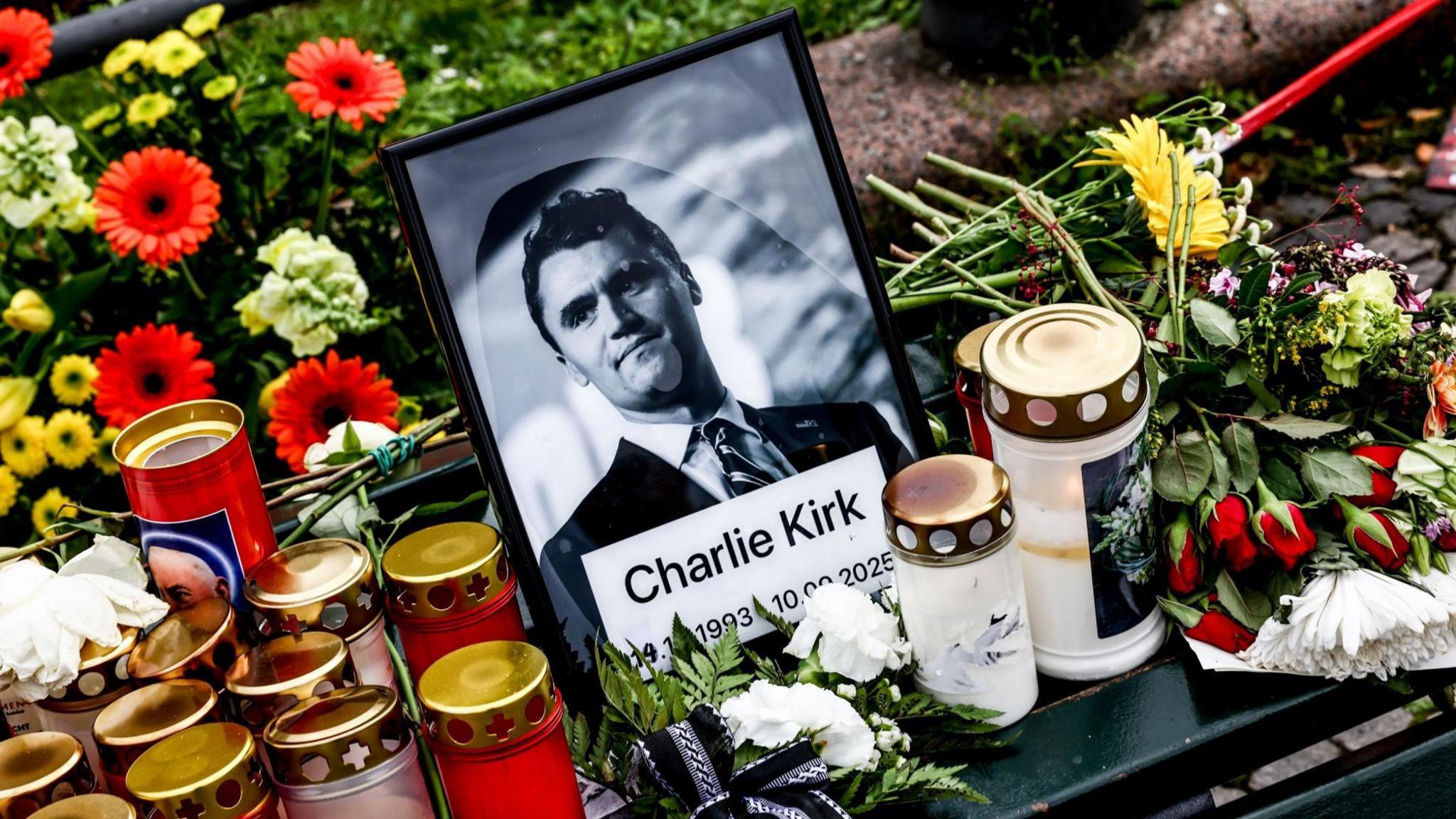 A black and white framed photo of Charlie Kirk, wearing a suit, is in the centre of a vigil. Next to the photo, there are candles and red and yellow flowers.