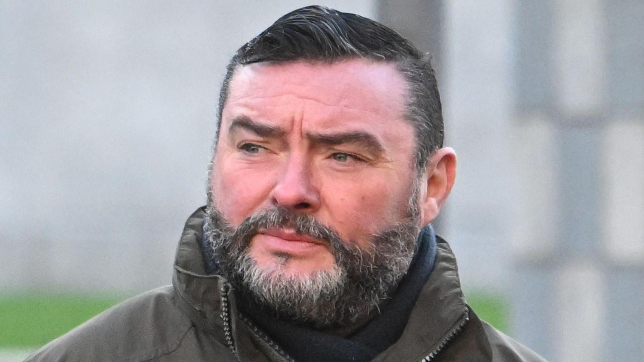 Shows a man with a bear, dark jacket and scarf