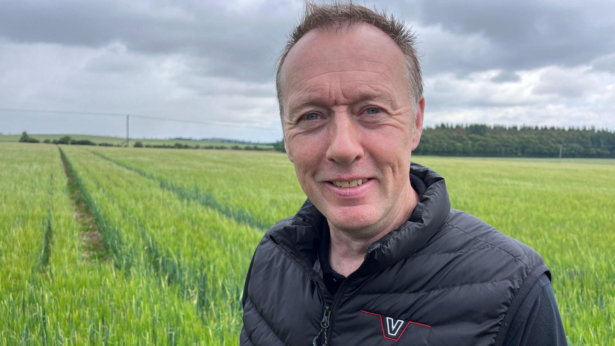 Neil White, with short, dark hair and wearing a black bodywarmer and tee-shirt, looks to the camera in front of his crop of spring barley.