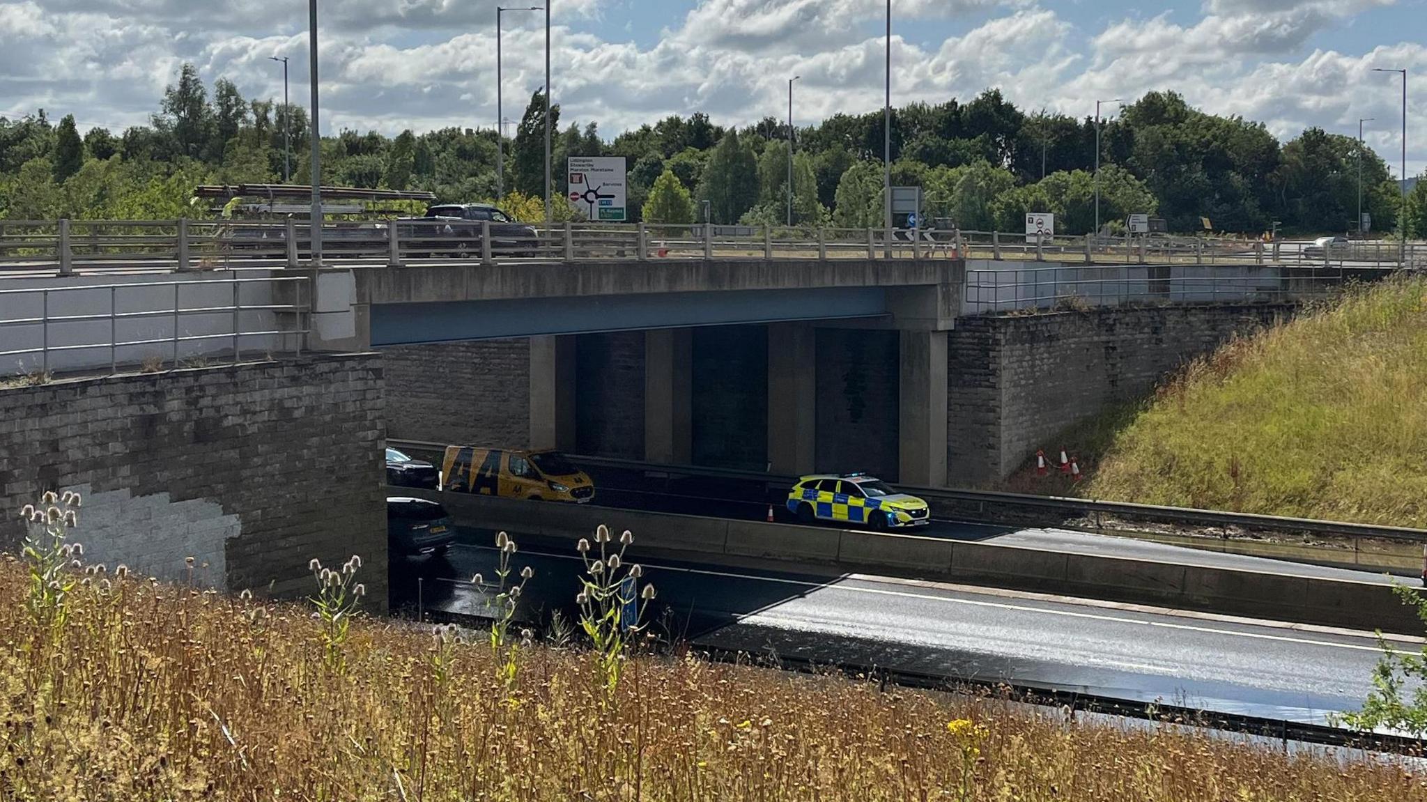 A police car in a coned off lane under bridge. There is flood water on both sides of the dual carriageway. In the background there are trees and a large road sign.