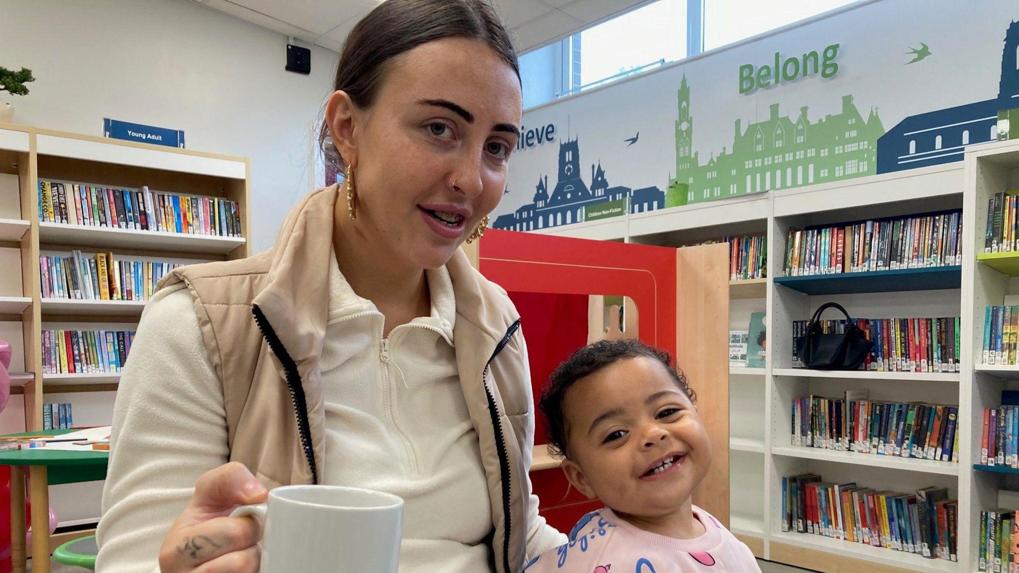 A woman with tied back dark hair holding a white mug, with her young daughter wearing a pink jumper and smiling.   