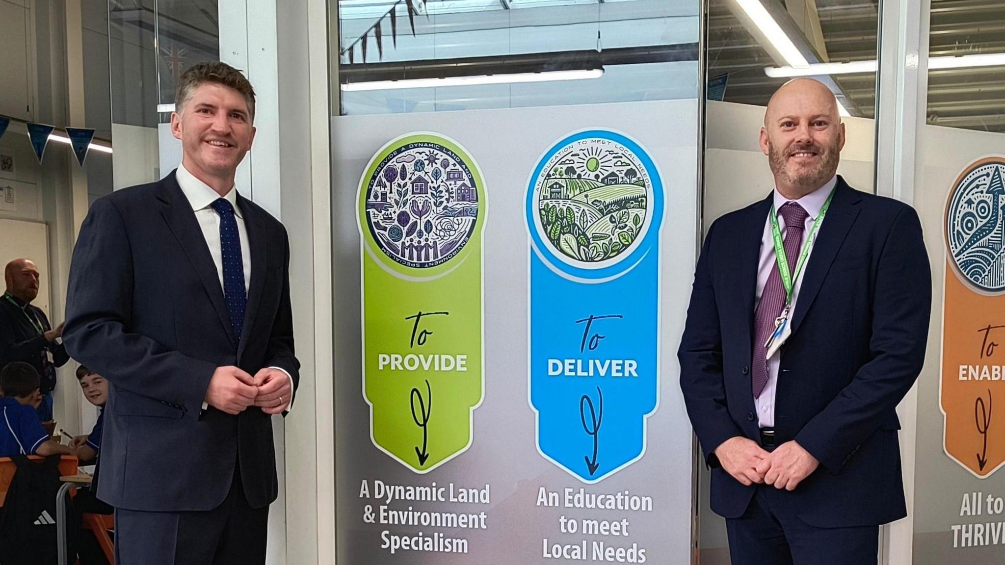 Edward Morello and Jason Malone smiling for the camera. They are both wearing dark suits and standing either side of a banner which says - to provide a dynamic land and environment specialism, to deliver an education to meet local needs.