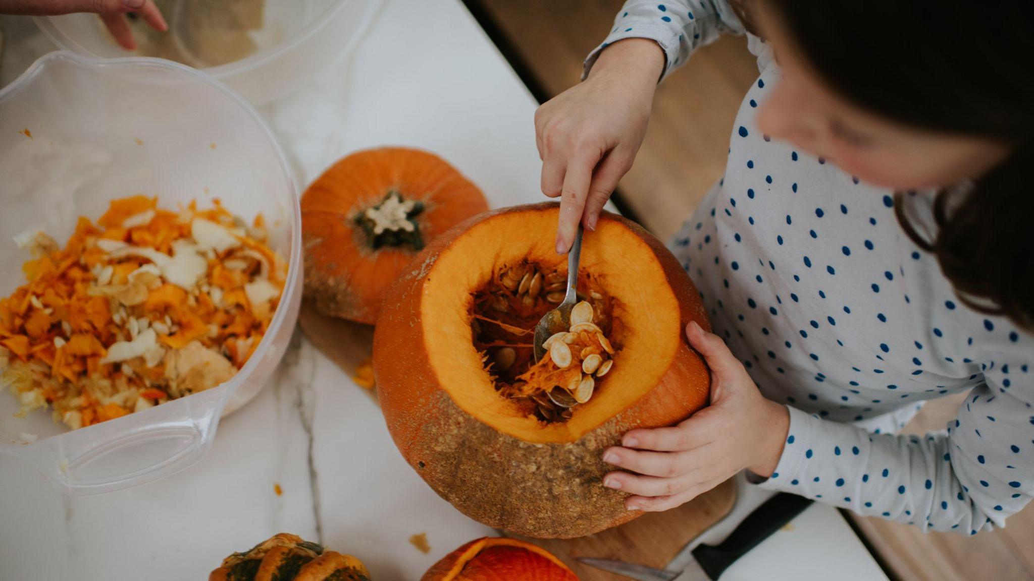 A stock image of a young girl scooping seeds out of a pumpkin using a spoon. 