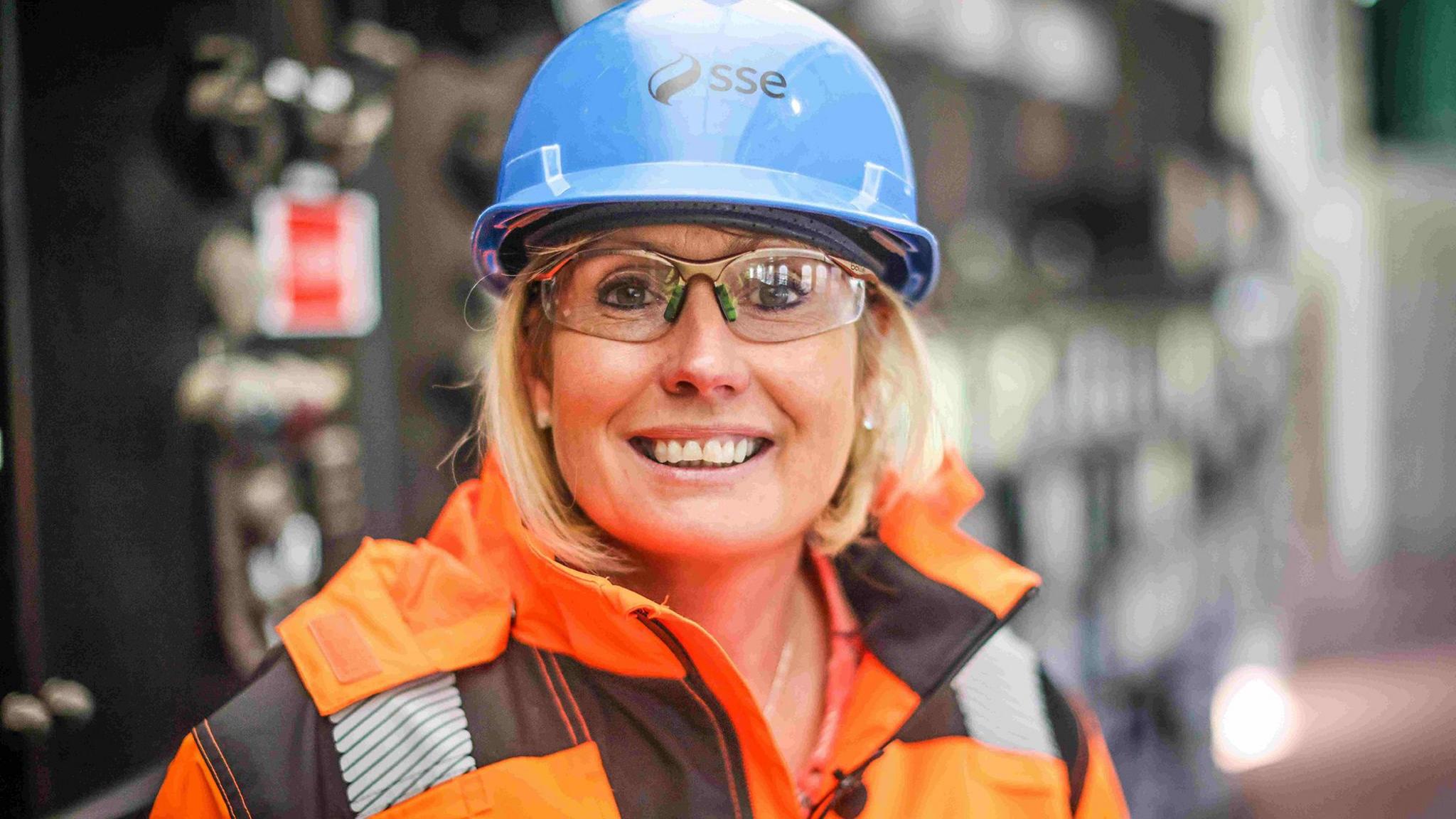 Gillian O'Reilly wearing hi-vis orange clothing and a blue hard hat and goggles stands in front of the control panel