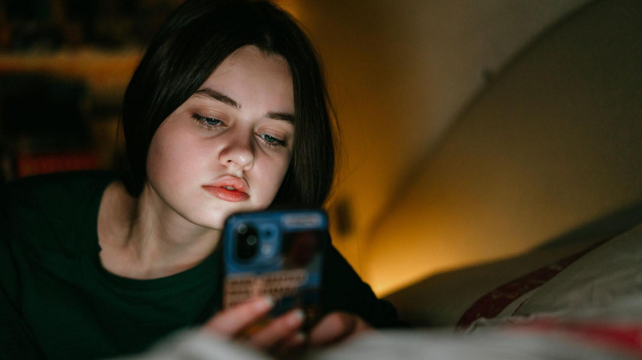 Portrait of a teenage girl looking at a mobile phone. She is laying on her bed. She has dark hair and a green jumper on and there is a light in the right of the photo.