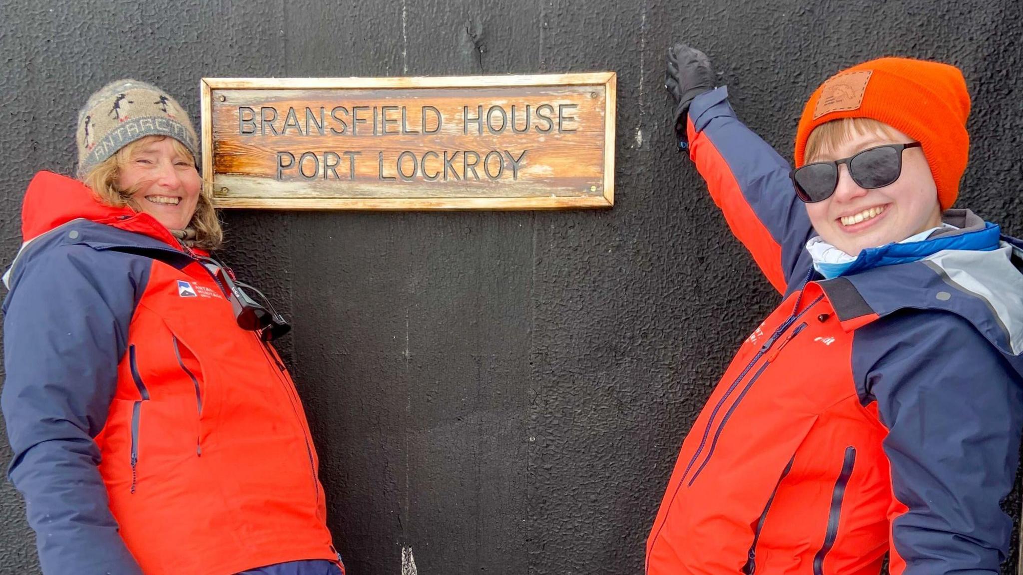 On the right is Aoife McKenna wearing  hat and glasses and a red coat and pointing at a sign saying "Bransfield House Port Lockroy". On the left is another team member wearing a red coat.