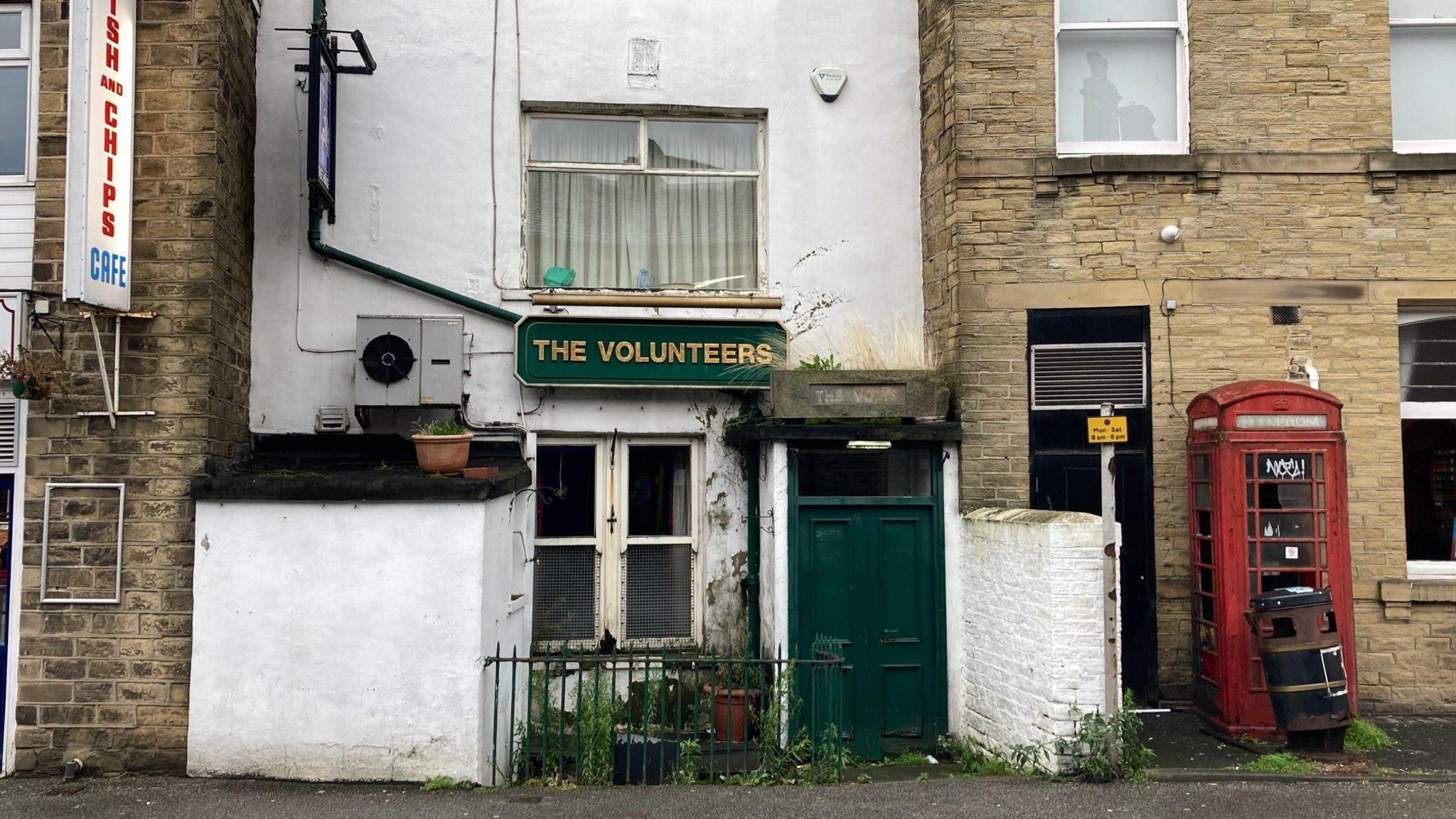 A whitewashed building with a green wooden front door squeezed in between two sandy-coloured, Yorkshire stone properties with a disused, red telephone box to the right of the entrance.