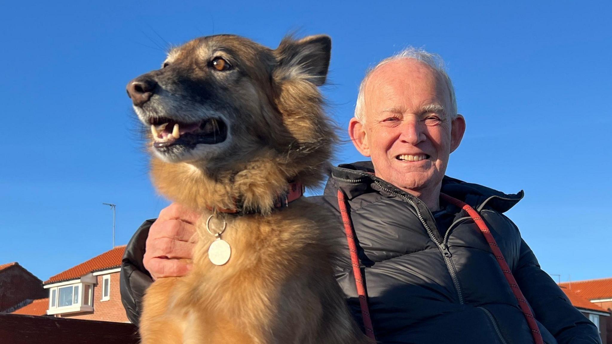 Cola sat on a bench with John Dalley who has his arm around the dog and a red lead draped round his black padded jacket