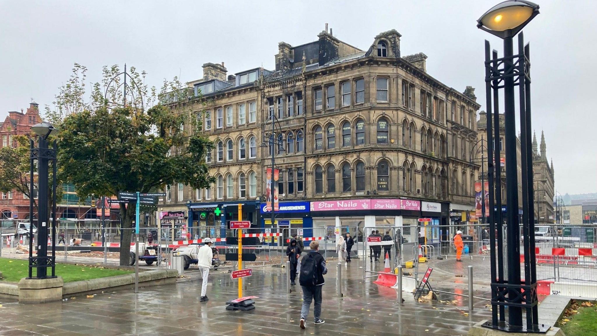 A five floor, Victorian building in Yorkshire stone with roadworks in the foreground on a wet pavement and a tree to the left hand side.  