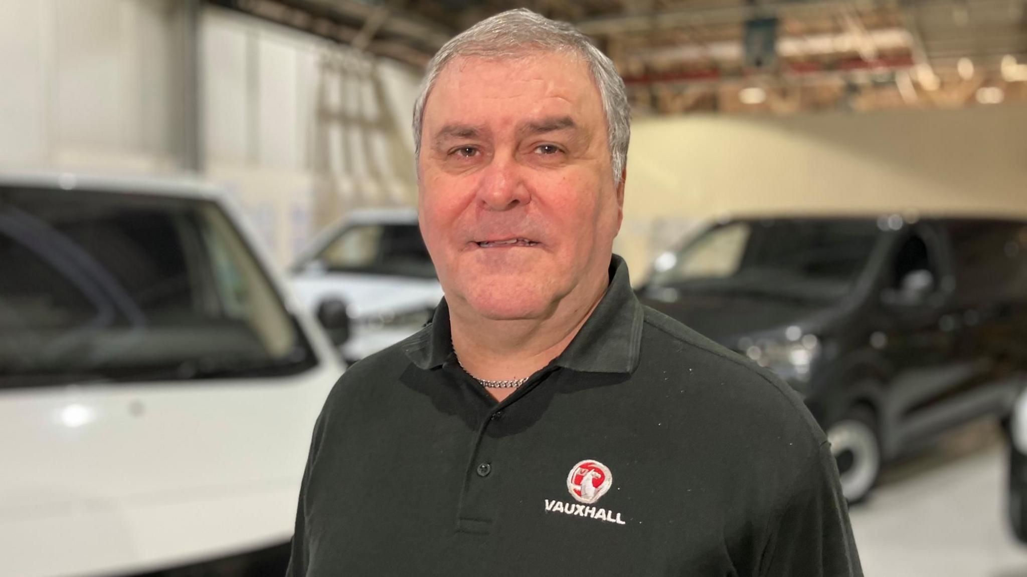 Mark Noble at the Luton Vauxhall plant wearing a black polo shirt with a red and silver Vauxhall logo. He has black and white vans behind him.