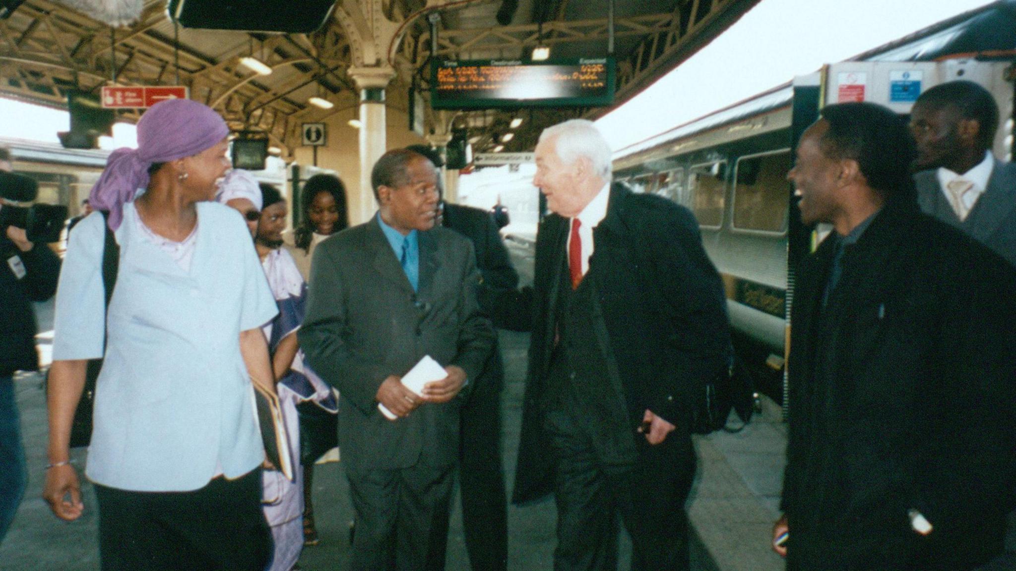 Dr Stephenson and Tony Benn walk down the platform of Bristol Temple Meads with a group of people. Mr Benn, who is wearing a dark coat and red tie, is smiling and turned to Dr Stephenson, and has his hand on his arm. Both are smiling at each other in greeting, and the people around them are walking and smiling at them too. 