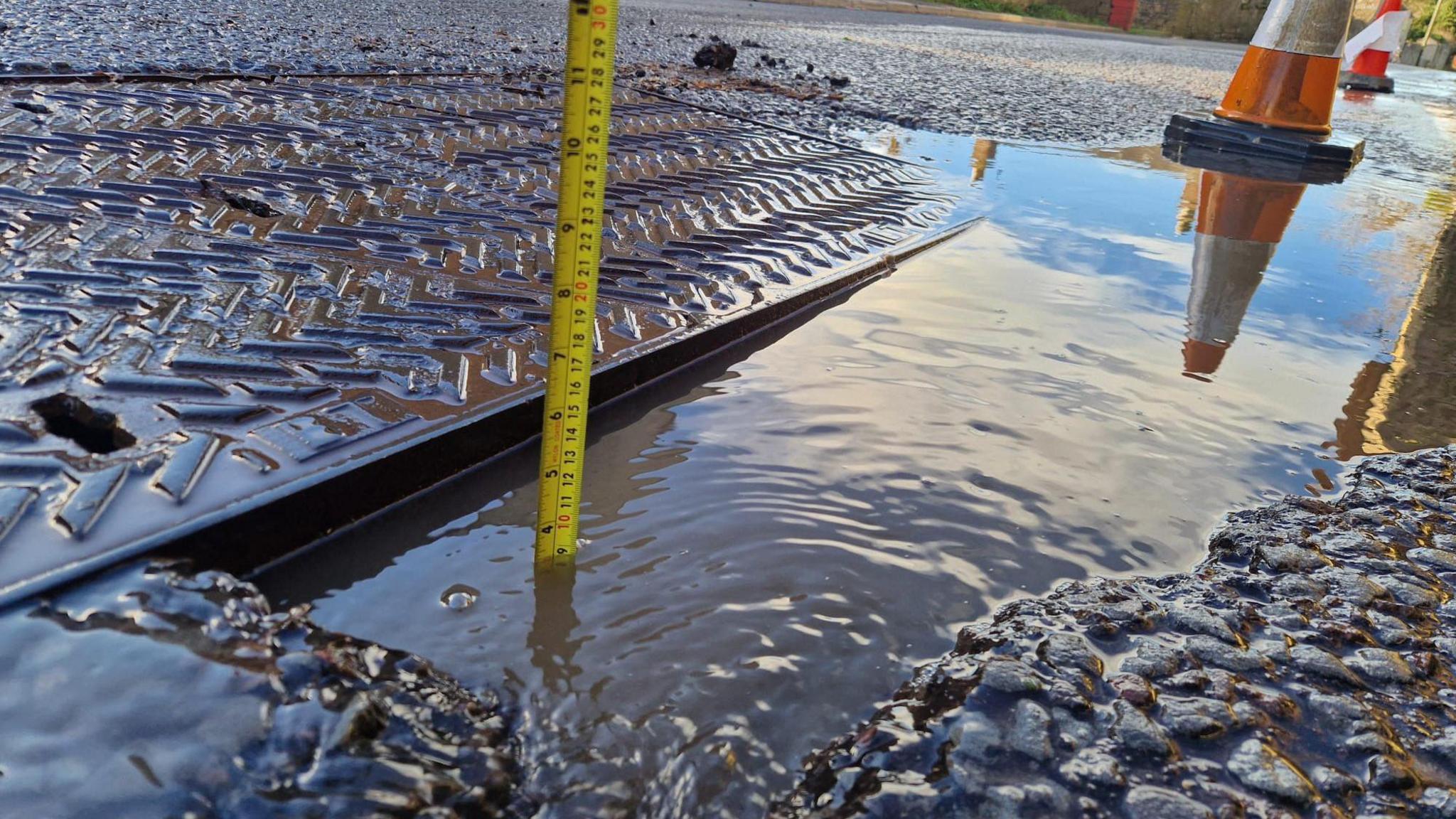 Yellow measuring tape being placed into the pothole  with a cone blocking part of the road