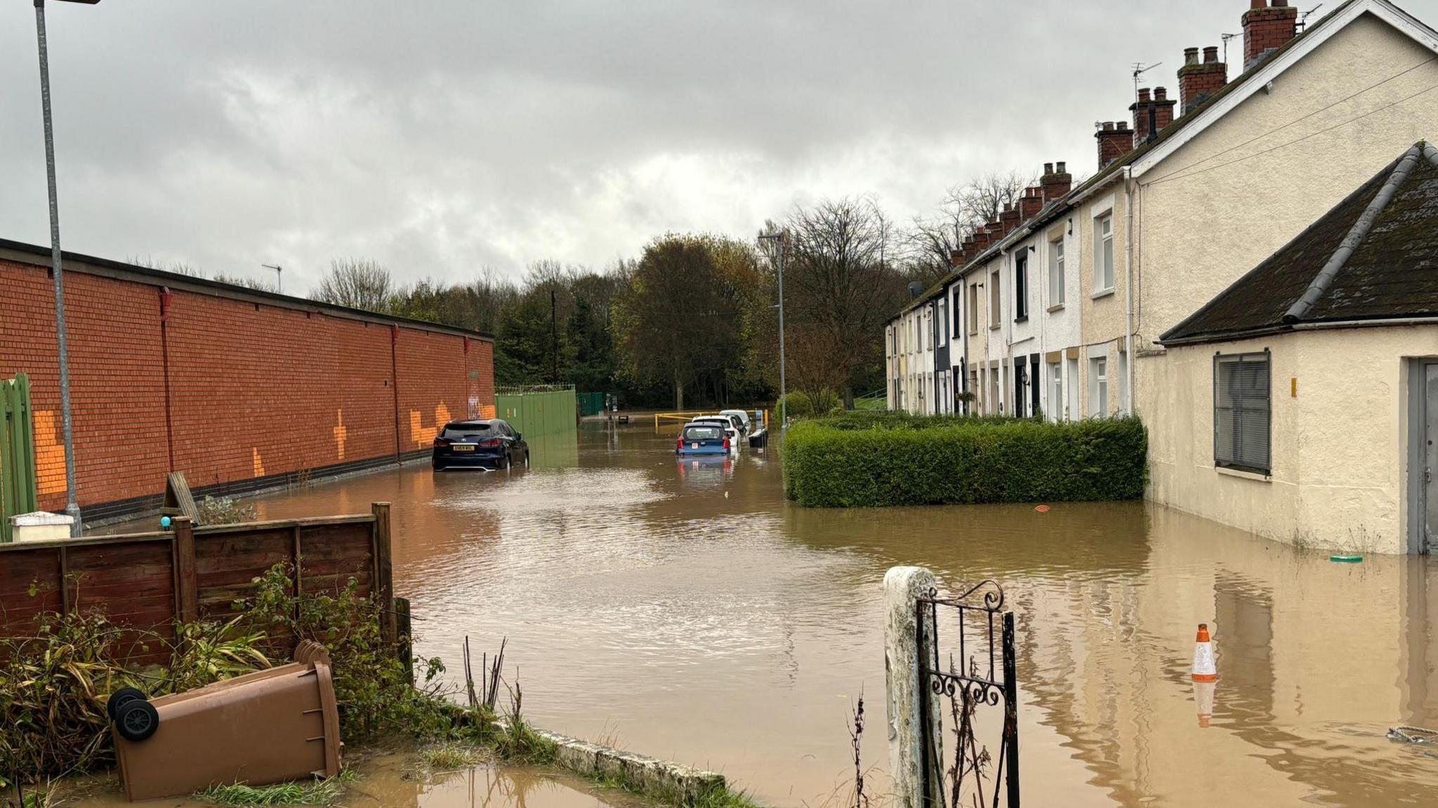 Flooding outside homes near Moat Park, Dundonald.  A row of terraced housing are surrounded by dirty brown water.  A number of parked cars are in the floodwater and a wheeled bin has overturned. 