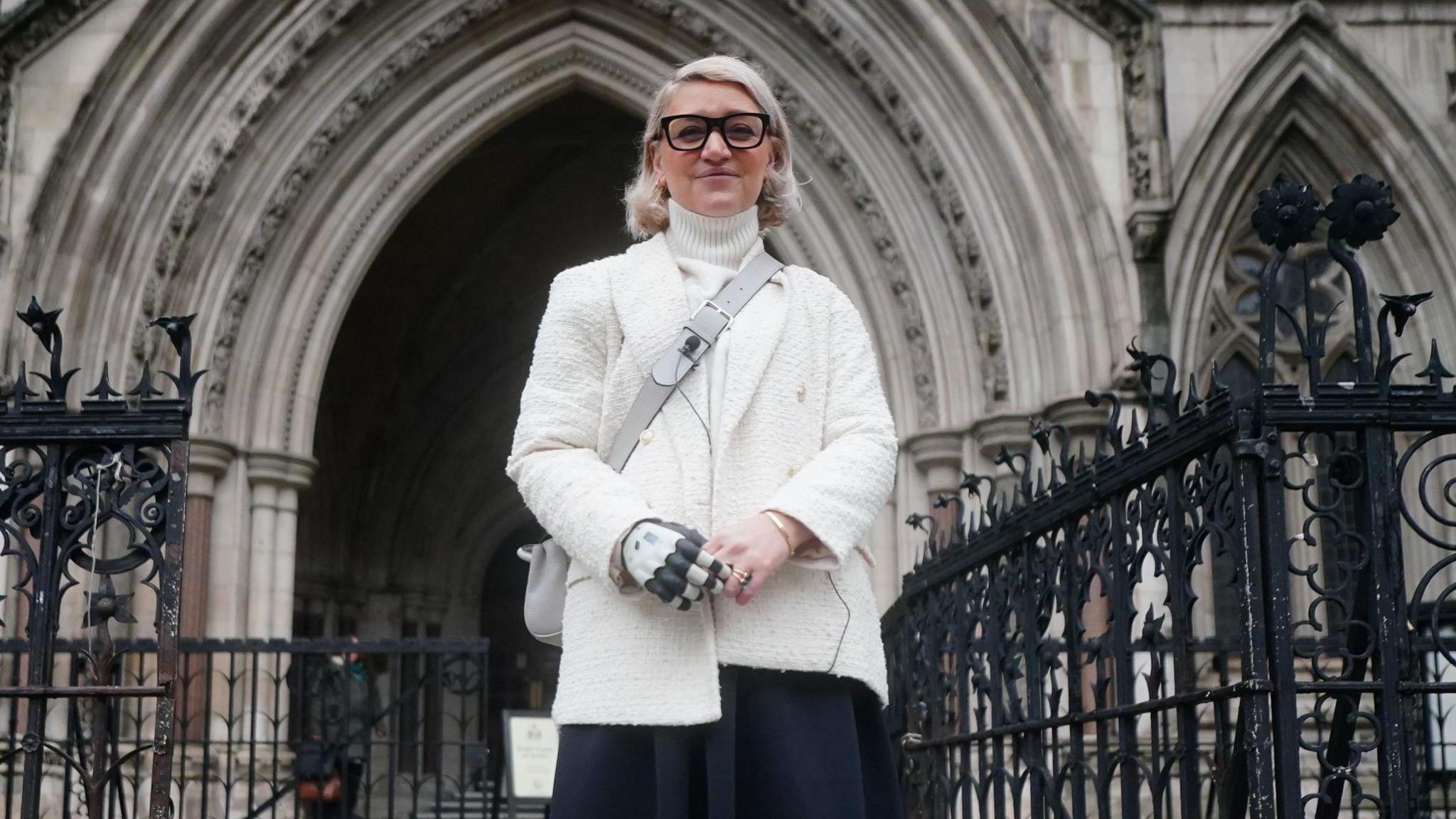 Sarah de Lagarde in a white blazer and black skirt stands in front of the Royal Courts of Justice. Ms de Lagarde has a white and grey bionic right arm. She wears a grey, crossbody handbag.