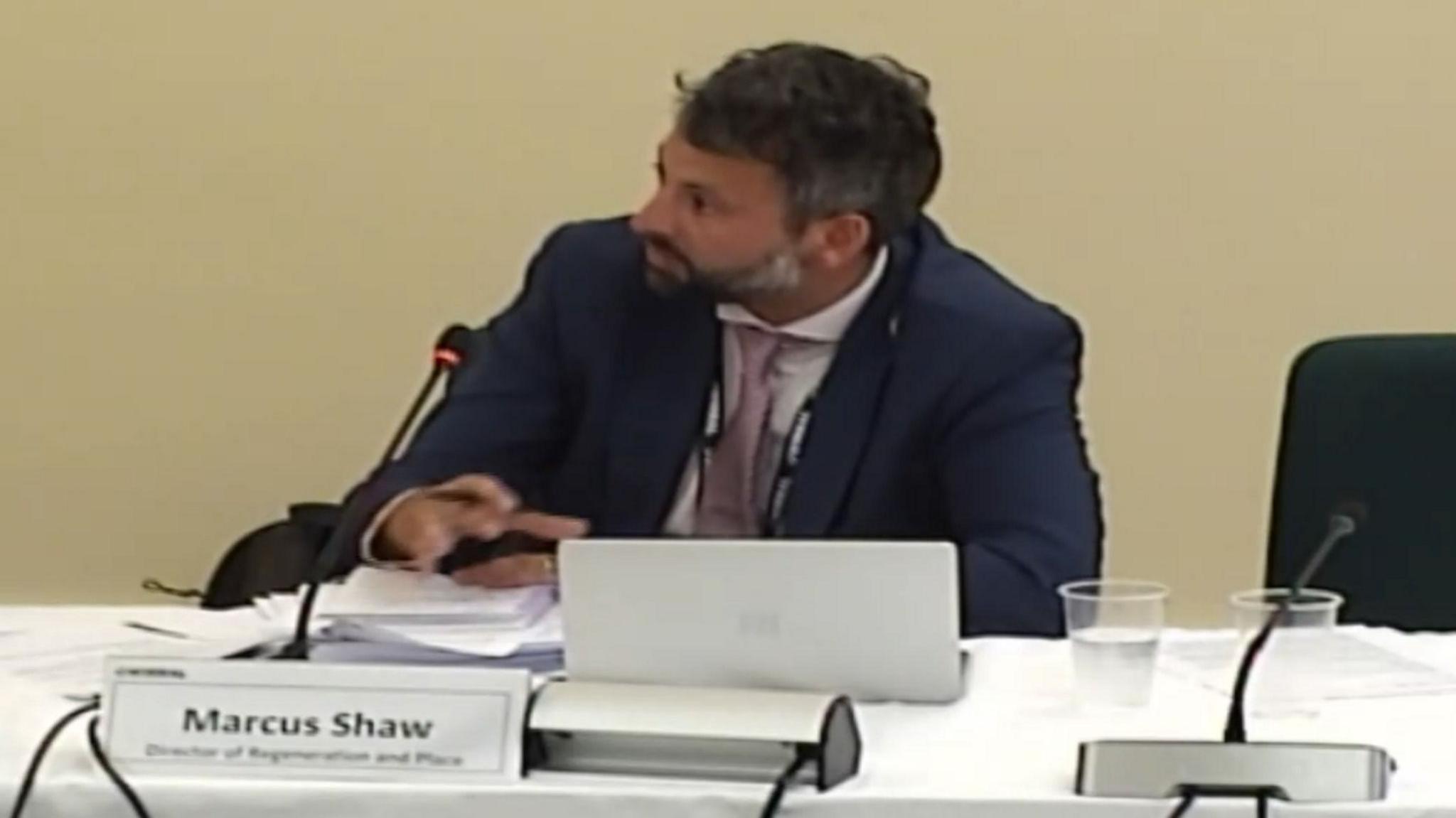 Marcus Shaw appears to be talking into a microphone while sitting at a desk during a council meeting. A sign in front of him includes his name and job title.
