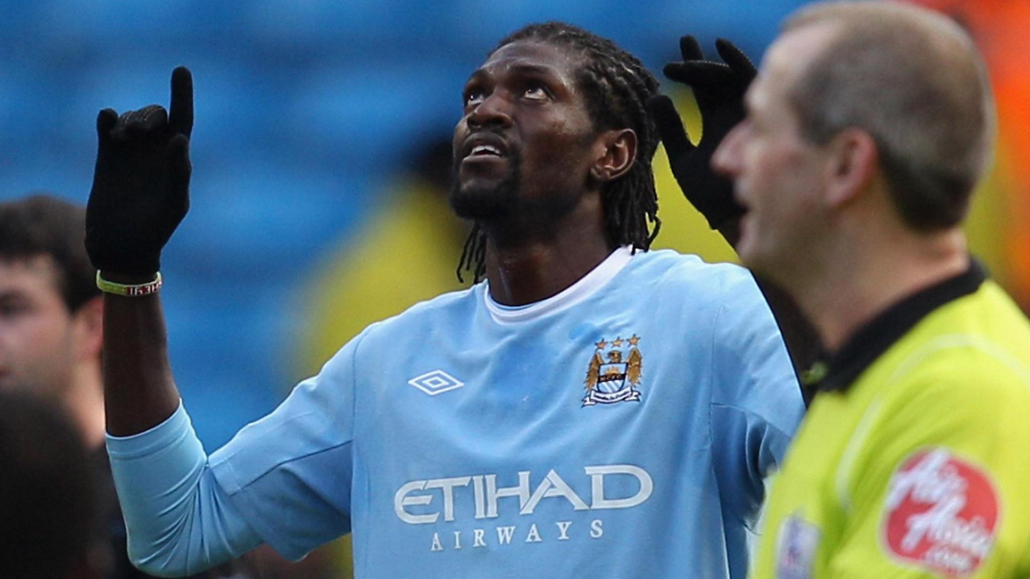 Emmanuel Adebayor, wearing a light blue Manchester City shirt and black gloves, looks up and points to the sky as he celebrates a goal, while a referee in a luminous shirt is seen out of focus in the foreground