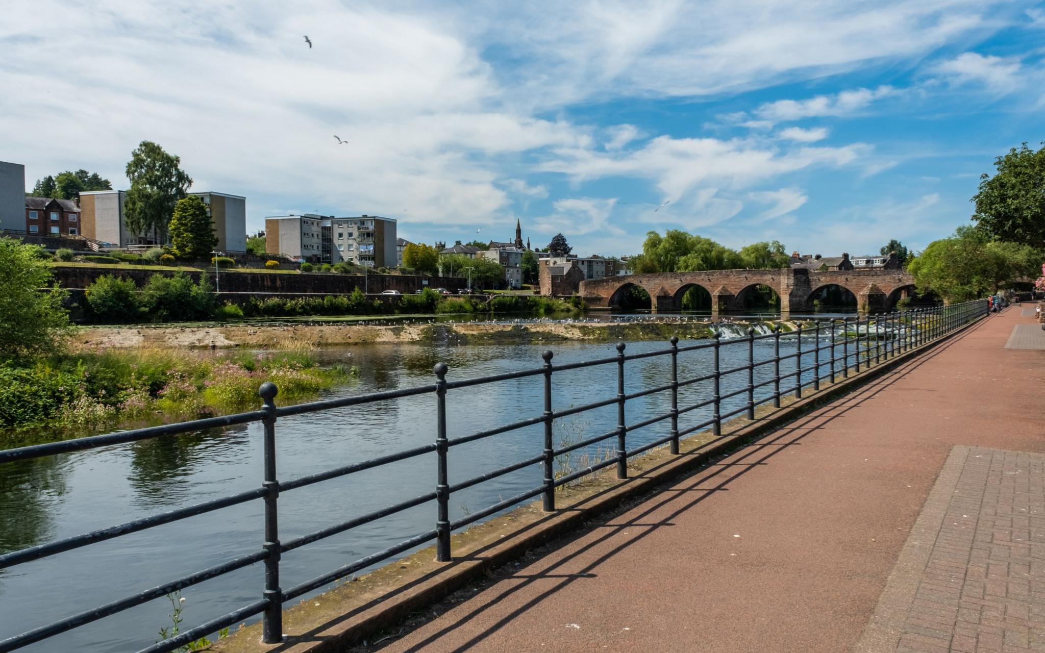 A view across a walkway to the River Nith in Dumfries. There is a sandstone bridge in the distance and blocks of flats on the other side of the river.