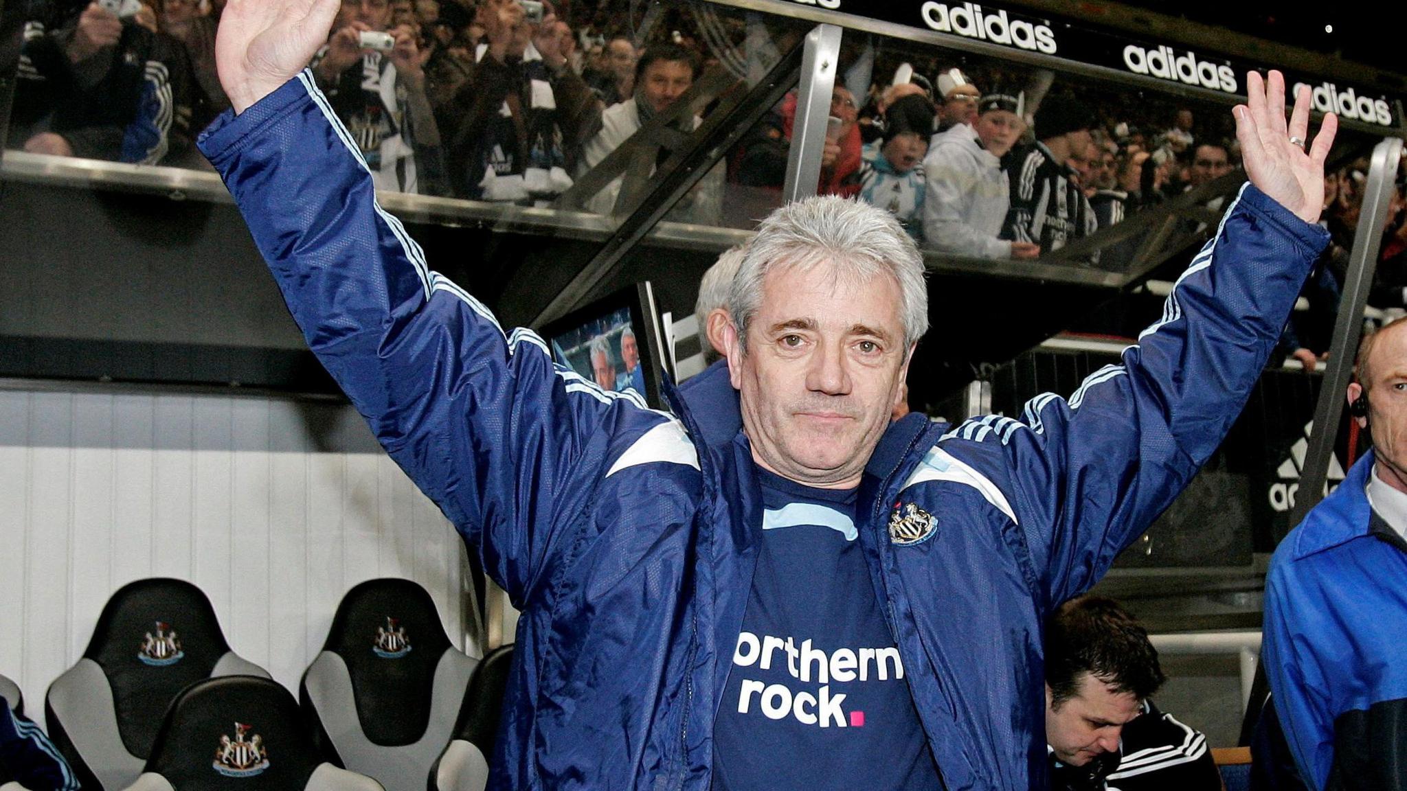 An old photo of Kevin Keegan when he was Newcastle manager in 2008. He has grey, short hair and is wearing a blue jacket with the Newcastle United logo and a blue top underneath with the Northern Rock sponsor peaking through. He is standing in front of the dugout at St James' Park and fans on the stands behind him. He has his hands in the air.