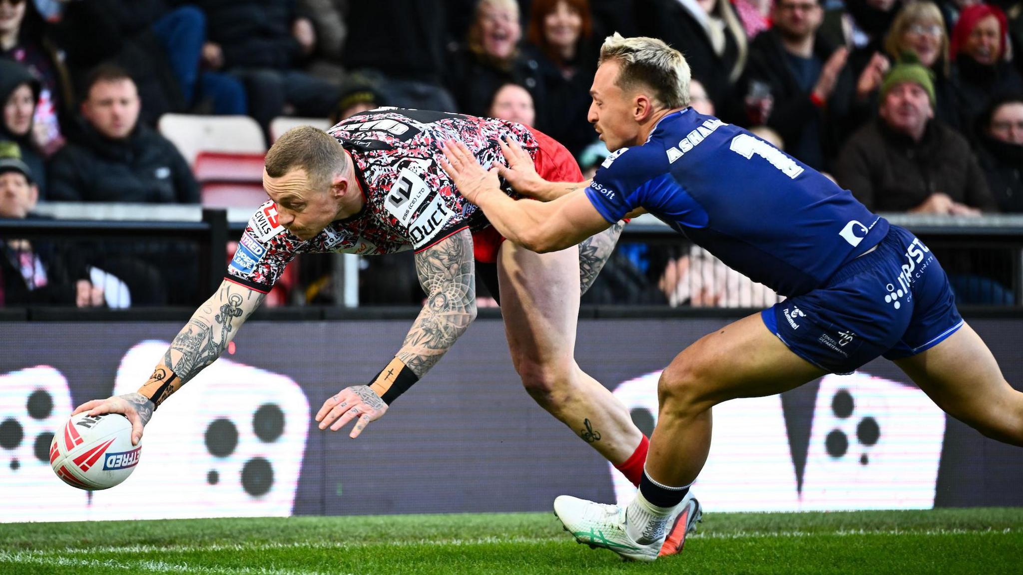 Josh Charnley with the ball in his right hand, about to touch the ball down in the corner as Toulouse full-back Olly Ashall-Bott puts two hands on his left side 
