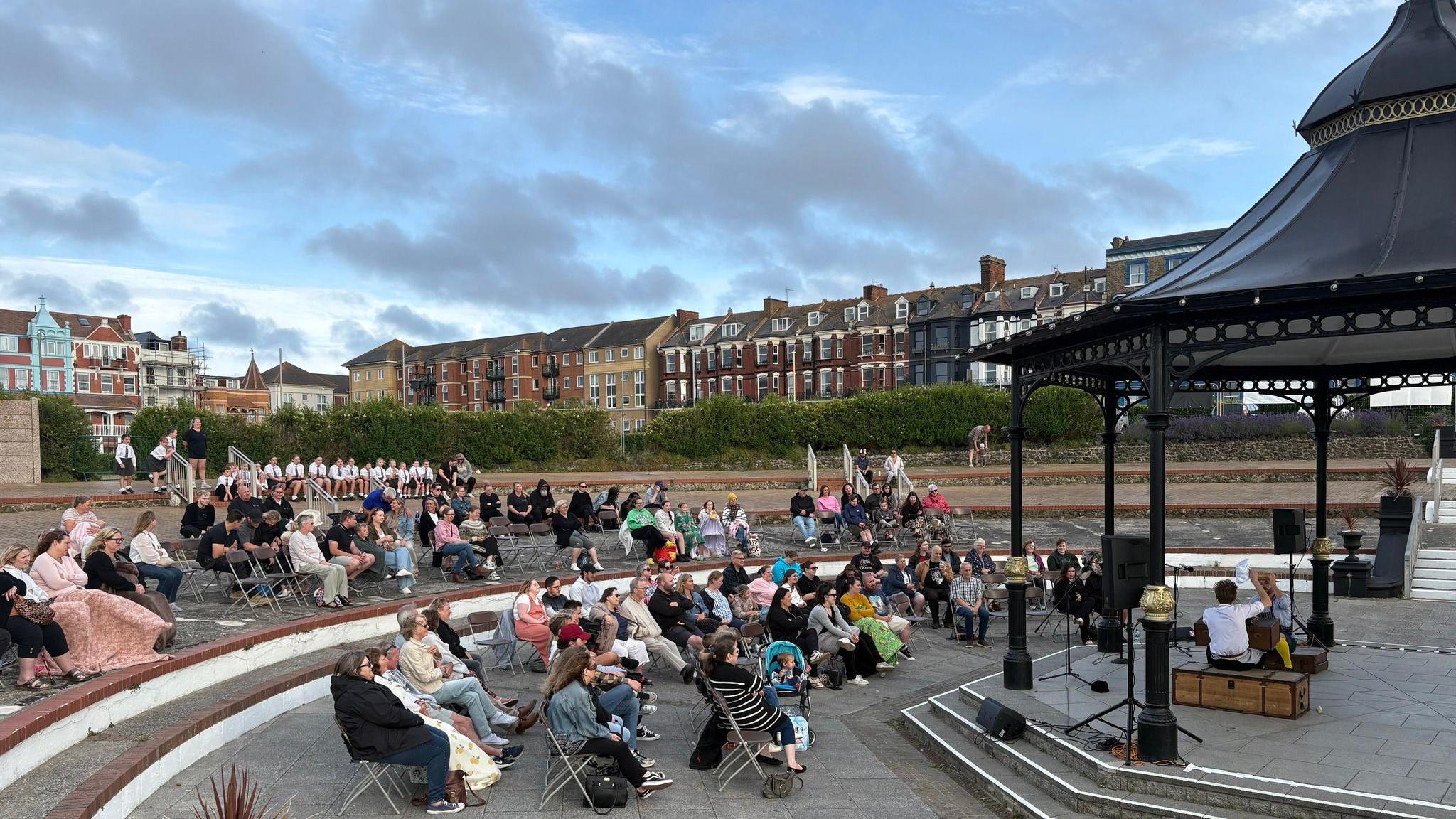 Dozens of people sat in chairs facing a black bandstand. There are green hedges and taller buildings in the background.