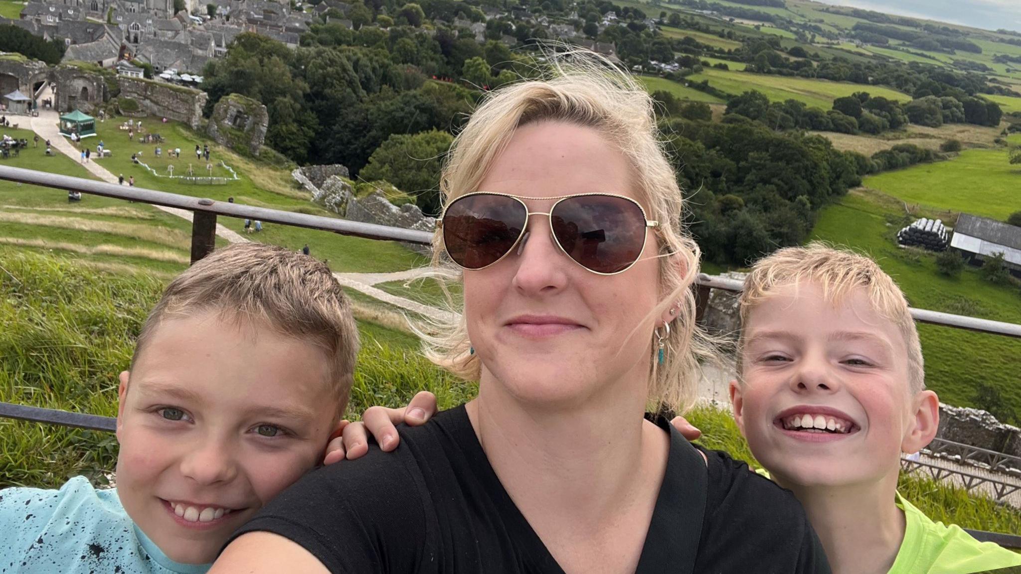 A selfie of Hayley Wyatt and her two children, posing in front of fields and trees. She is wearing a black t-shirt, has blonde hair tied up in a ponytail and is wearing dark sunglasses. Her two young boys are standing beside her with their hands on her shoulders, grinning at the camera.