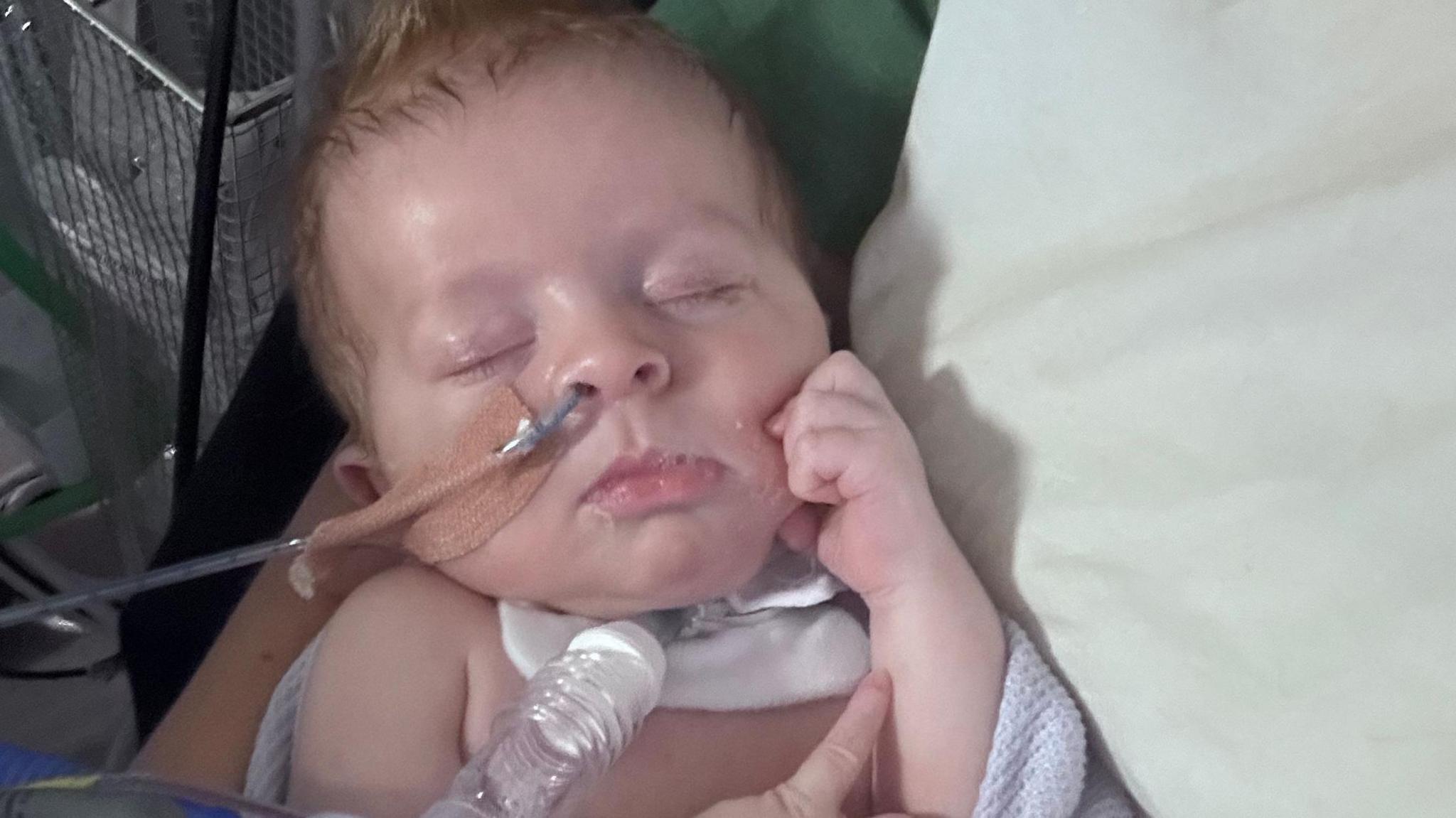 Baby Joshua is seen sleeping with his eyes closed, lying in his parent's arms. Joshua has light hair which is spiked at the top of his head. He has a tube into his nostril, secured with a nude plaster. He also has a white tube, secured with a white bandage onto his neck. 