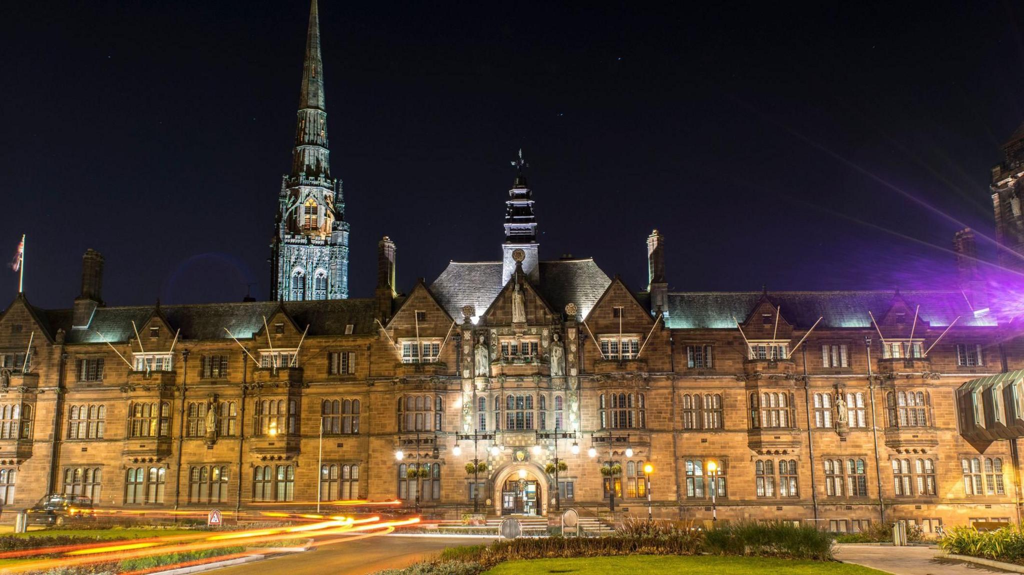 Coventry City Council building illuminated at night. The stone building has a number of windows with lights on. Behind the council building is a spire which is lit up against the night sky.