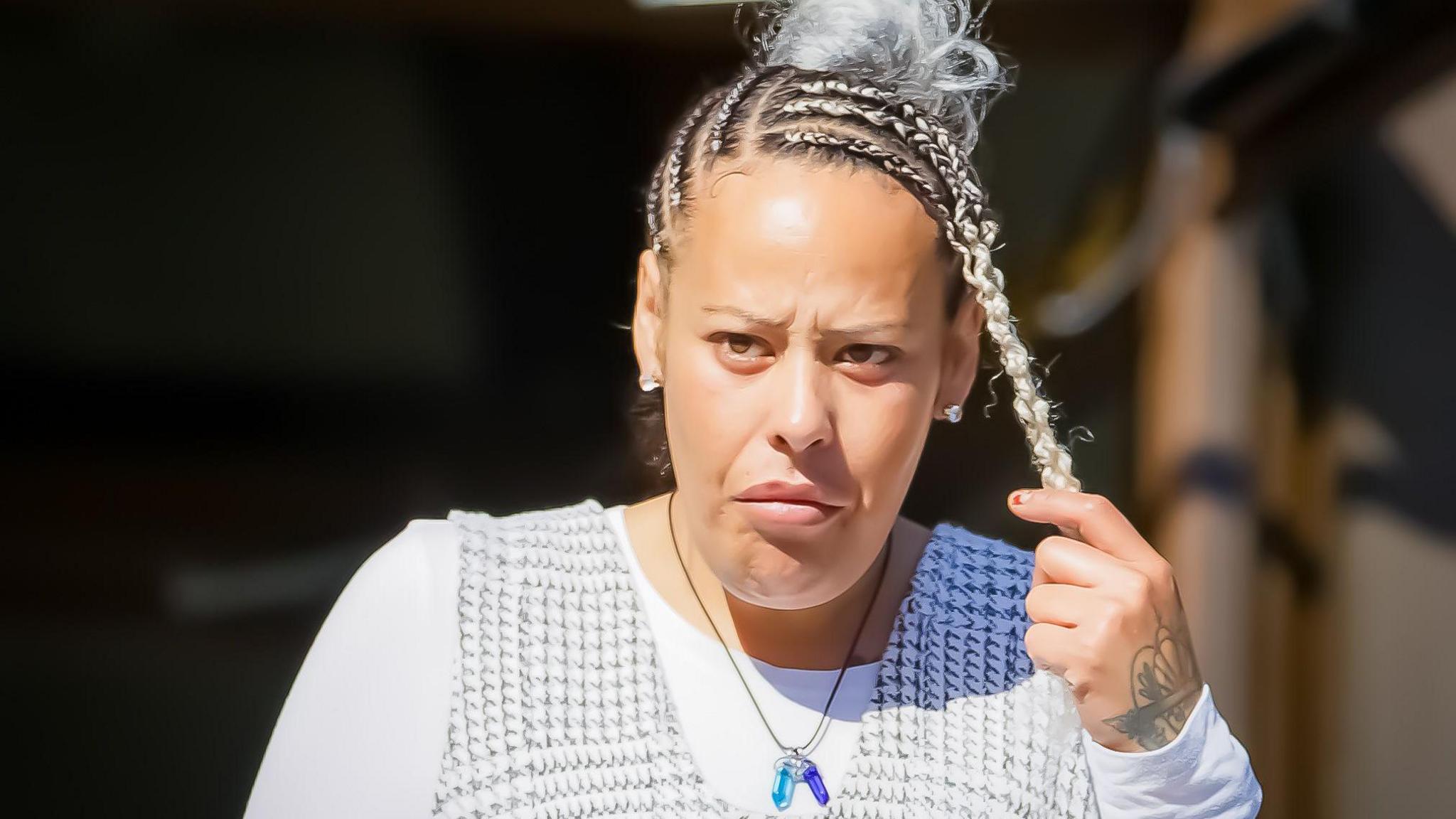 A woman stands outside the courthouse looking to the left of the photographer. she's has her hair in braids tied on top of her head and is wearing a black check dress over a white shirt. she is holding one braid in her left hand revealing a tattoo on the back of her hand.