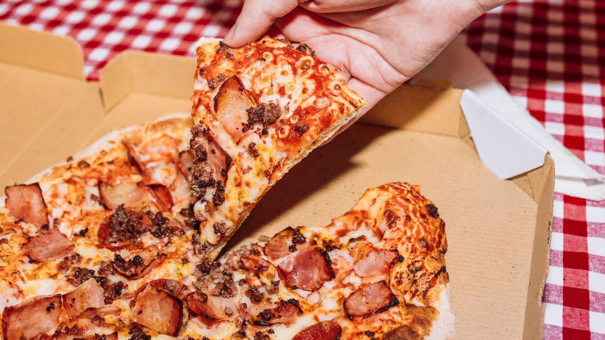 A hand picking up a slide of ham pizza from a takeaway box on a red and white gingham tablecloth.
