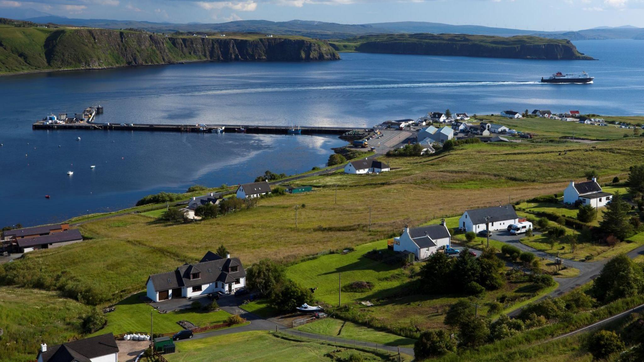 White-walled houses and other properties in Uig on Skye. The settlement is on the shores of the sea. There is a Caledonian MacBrayne ferry sailing off from Uig harbour. The land is green and the photo was taken on a fine, bright day.