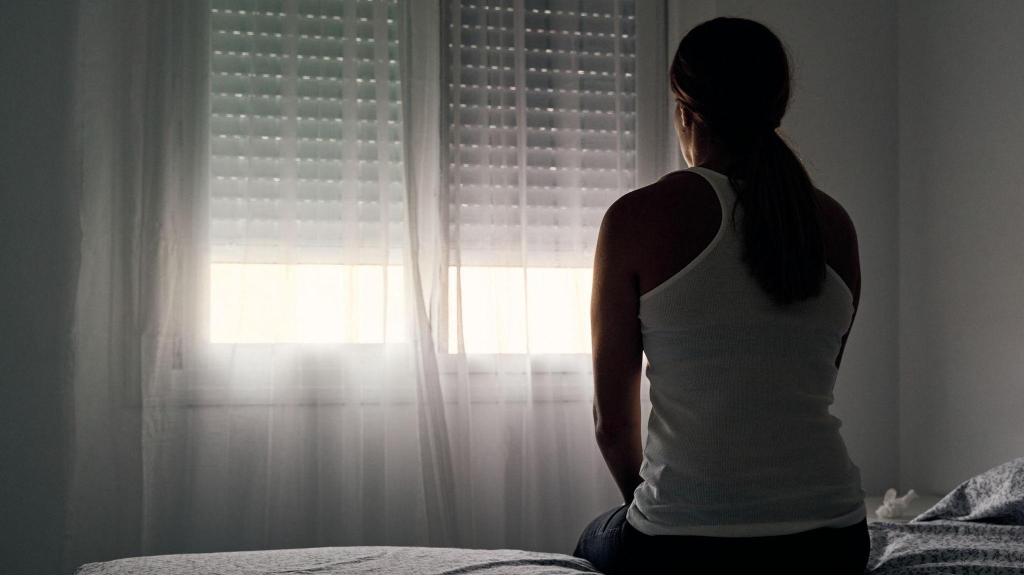 A woman with a long dark pony tail sits on a bed facing a window with the curtains and blind closed. She is wearing a white vest top. There is daylight coming through the lower part of the window and the walls are white.