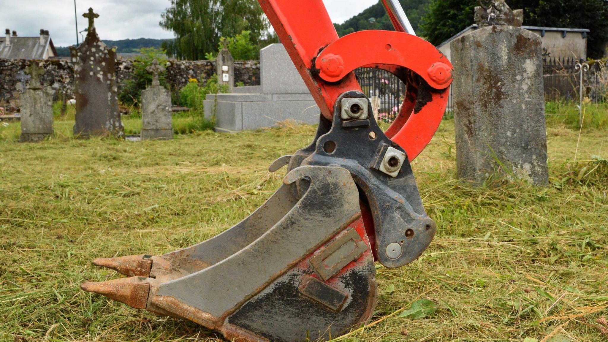 An excavator bucket with a red arm in a cemetery. The metal bucket is worn and rusty. In the background are old grave stones, some weathered and covered in moss. A stone wall and trees surround the cemetery. The grass is long and green. 