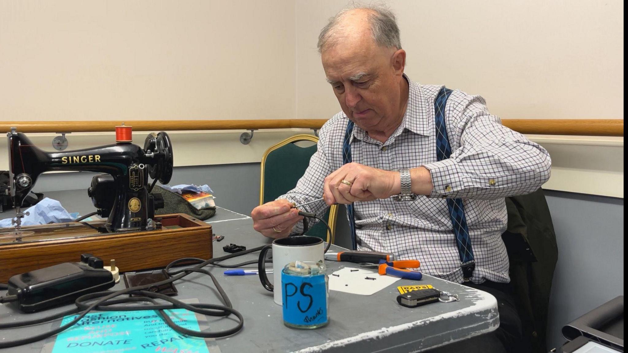 A man with braces and a checked shirt fixing a Singer sewing machine with a small screwdriver.