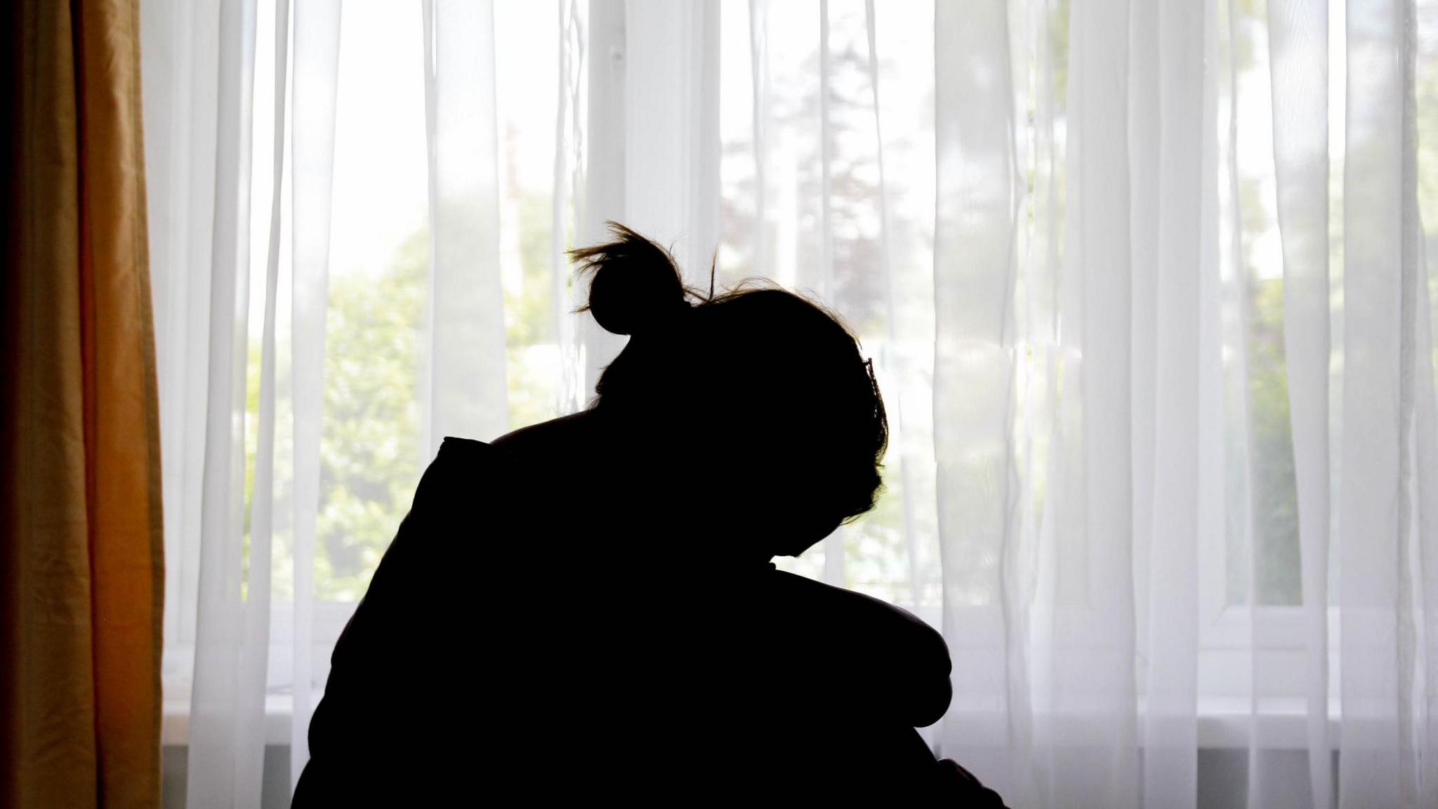 A silhouette of a woman in front of a window, which has a curtain drawn across it.