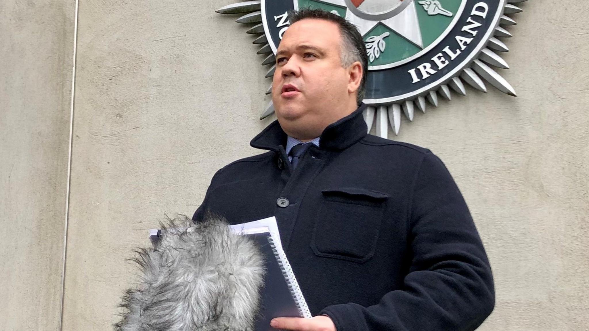 John Caldwell speaking at some microphones holding a notepad and standing in front of a PSNI logo