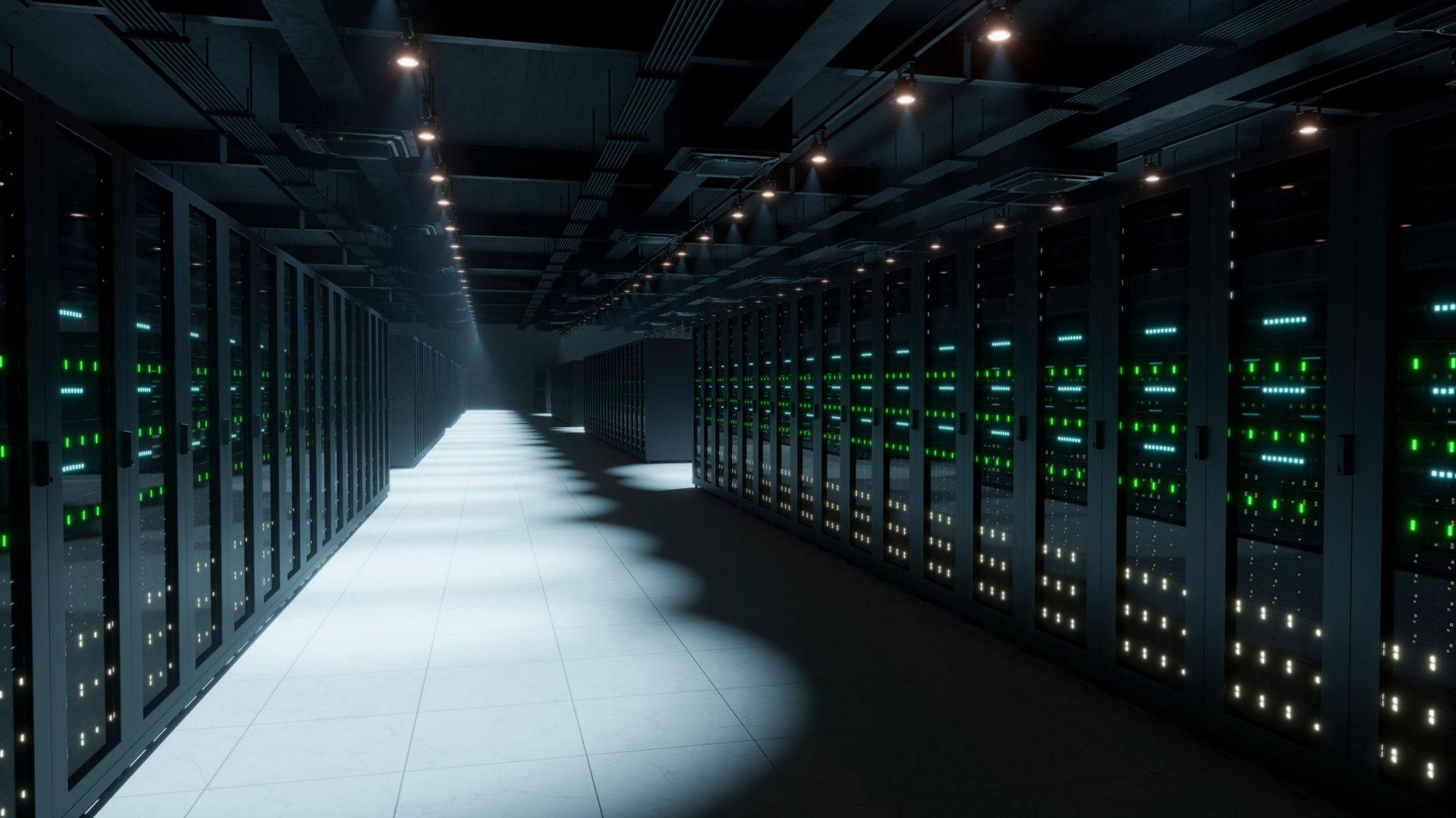 Rows of computer servers with LED lights fill a dark room. The floor tiles and some of the systems are illuminated by spotlighting.