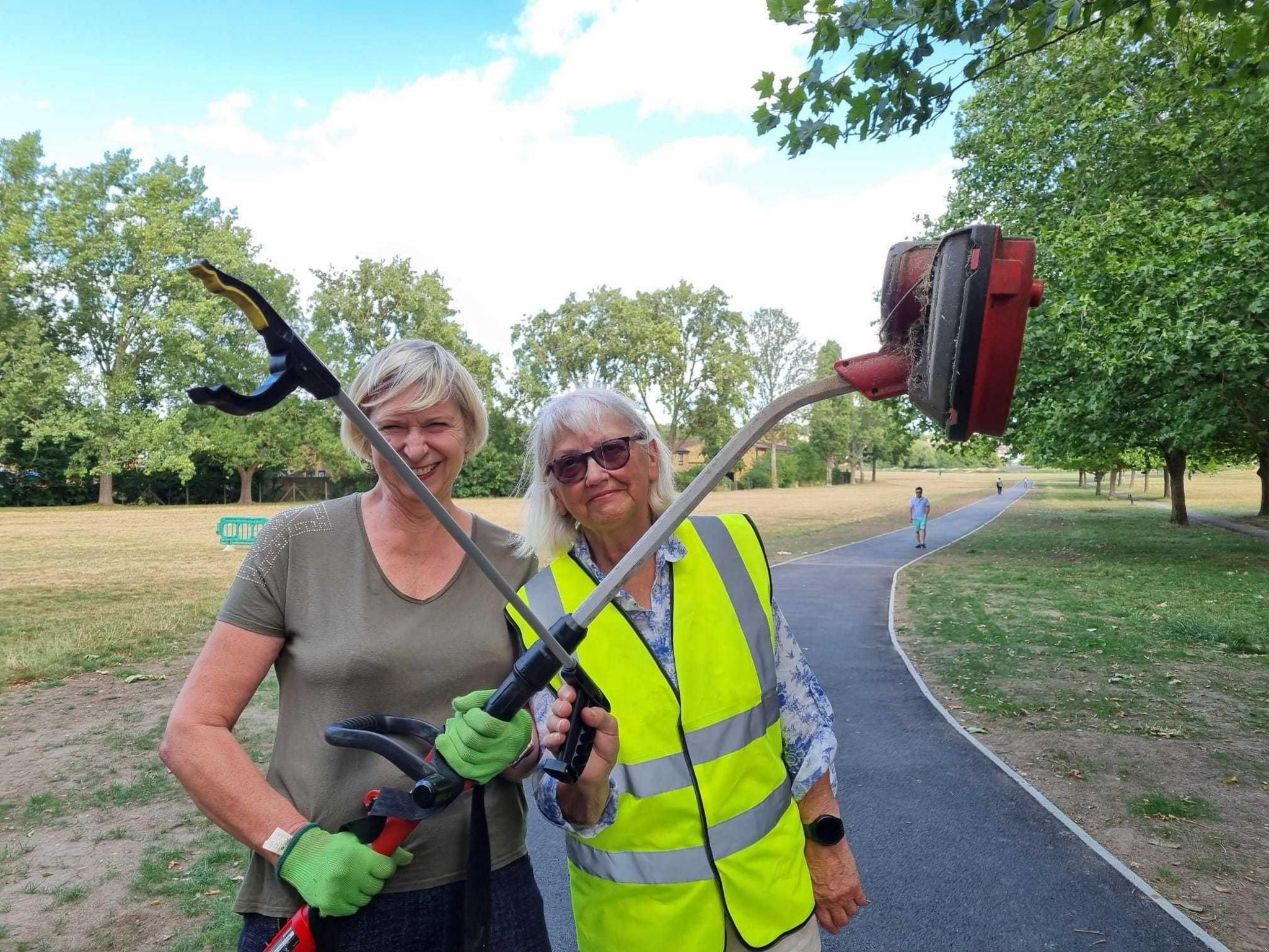 Two women are standing in the middle, holding two large implements used for litter clearance. On the left, a woman with short blonde hair is wearing a brown-green T-shirt and bright green gloves, and is holding a red sweeper in both hands. On the right, a woman with mid-length grey hair is wearing sunglasses, a white shirt with blue floral decorations and a bright yellow Hi Vis vest, and is holding a litter grabber in her right hand. They are standing on a grey tarmac path, which is running through a field of yellow-green grass, with trees a short distance away on either side.