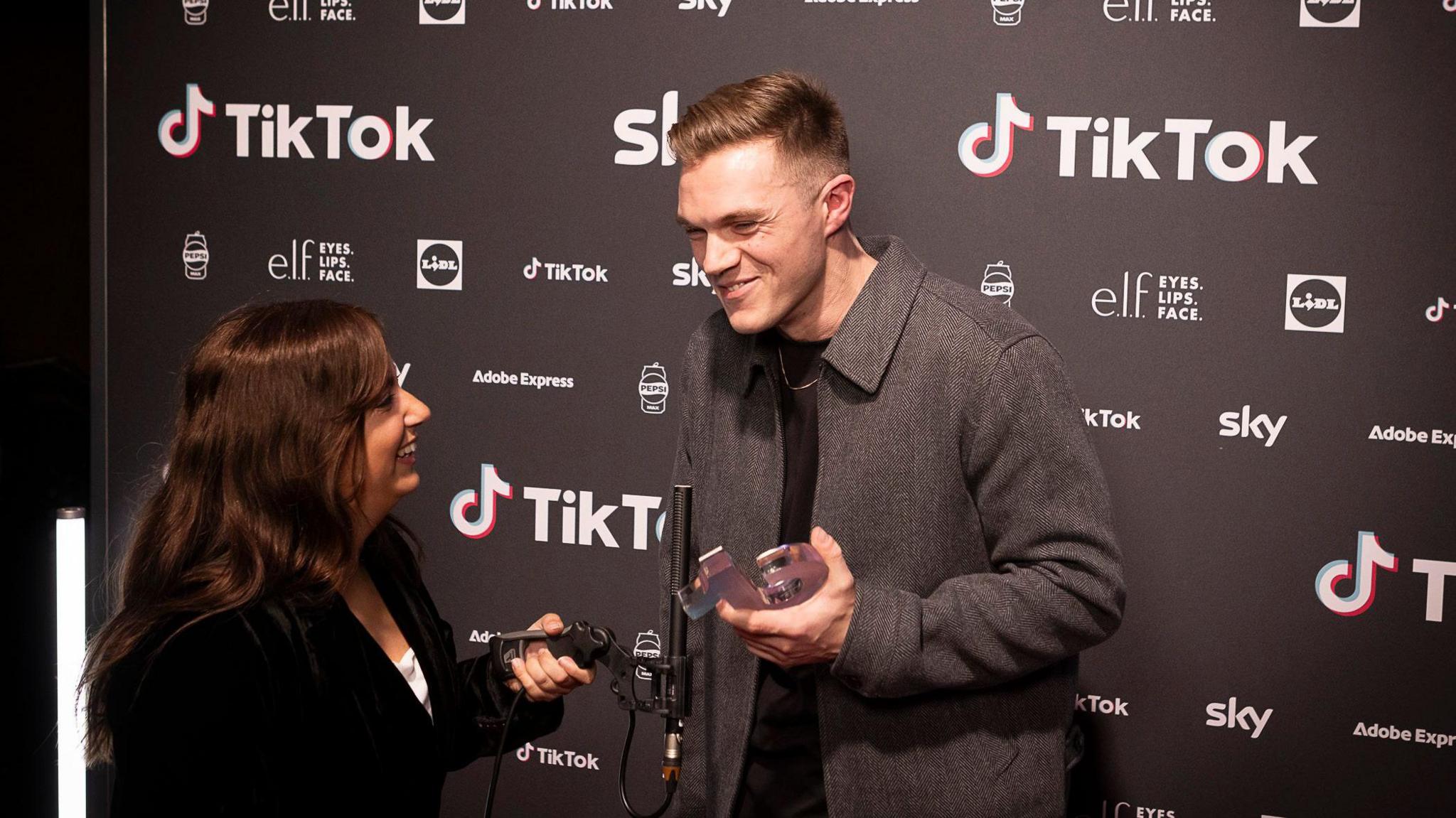 A man and a woman stand laughing together. The man is holding an award and is stood to a black and white backdrop with the words TikTok on it.