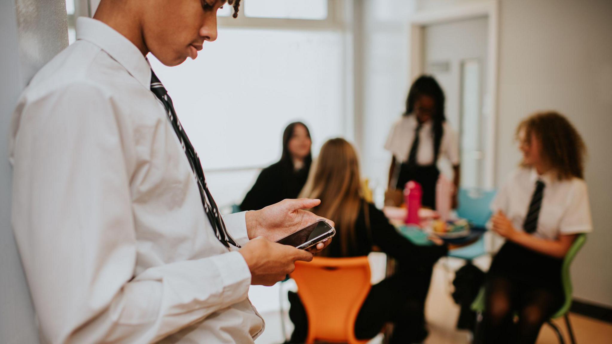 A boy in school uniform stands with pupils around a table in the background. He is on his phone