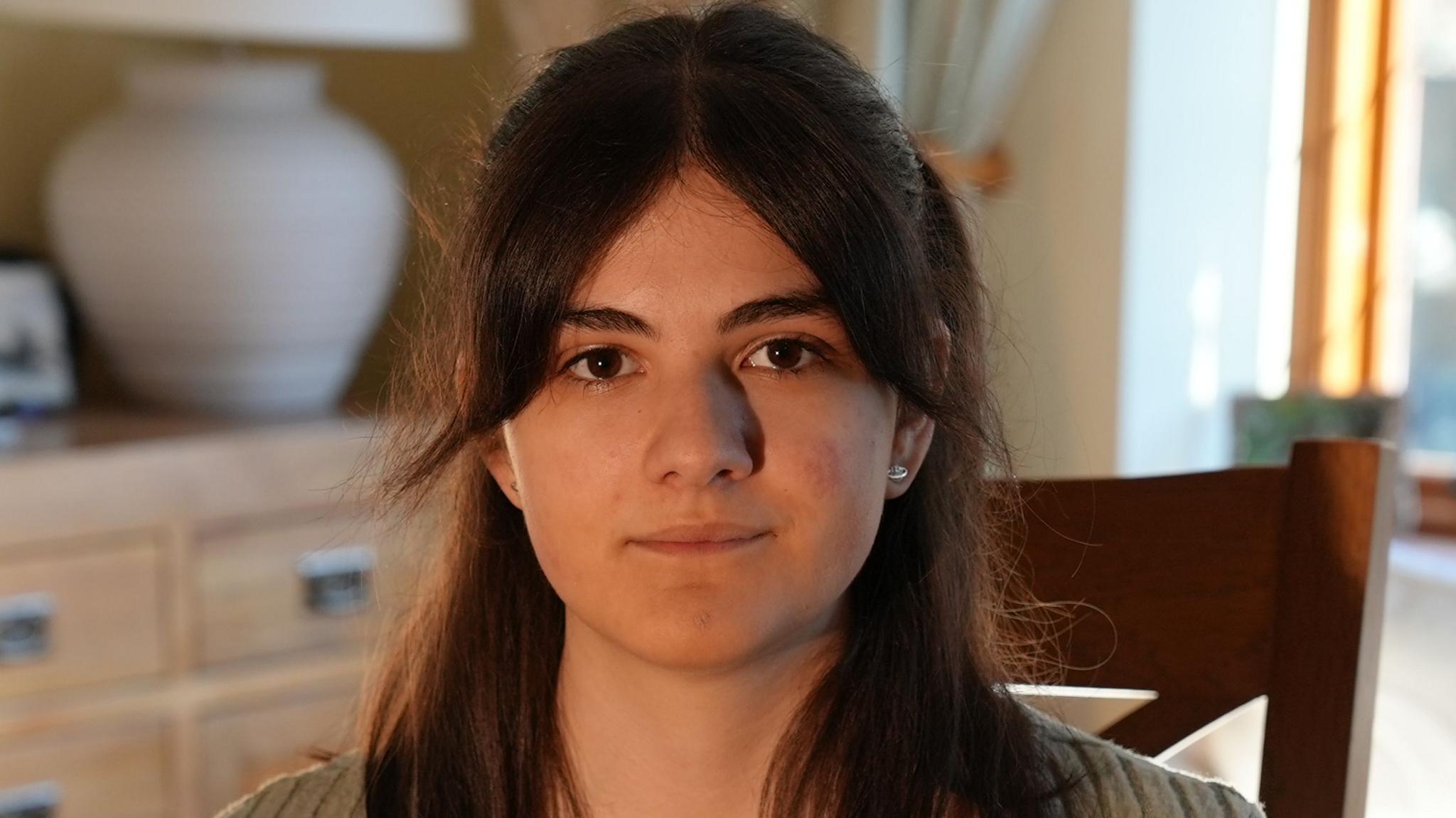 Kaylee, who has brown shoulder length hair and a fringe, wearing a green top and necklace, looks at the camera