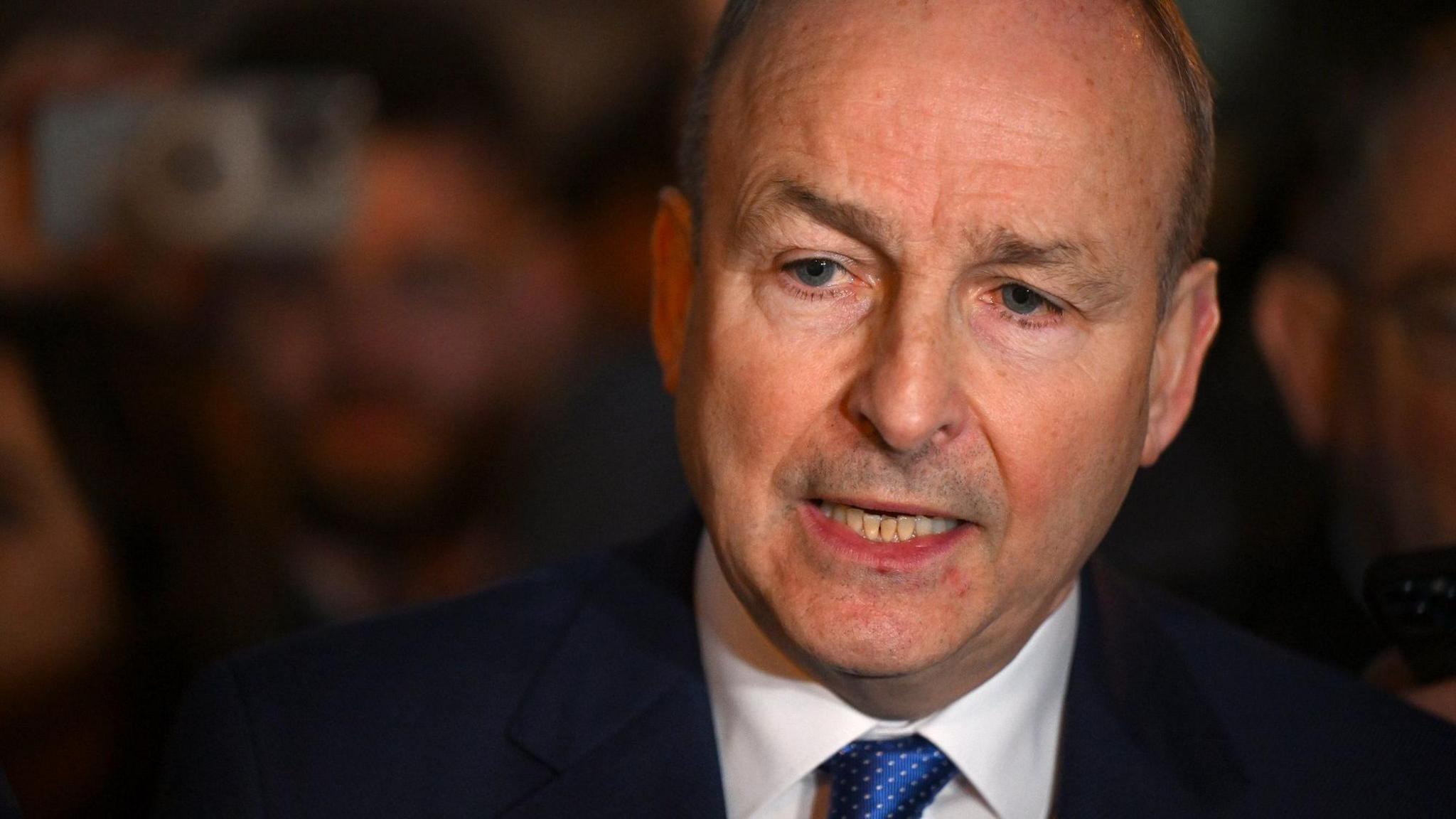 A close-up photo of Micheál Martin. He has short, receding brown hair and is wearing a dark suit with a white shirt and a blue polka dot tie