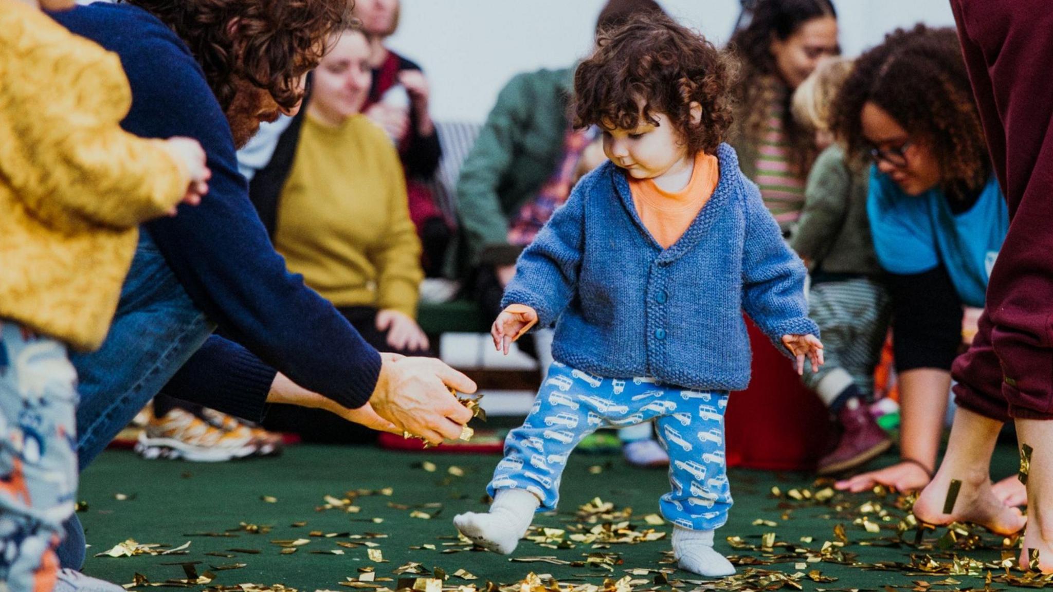 A toddler at one of the sessions hosted by Travelling Light theatre company. He is walking along, surrounded by a group of adult and other children who are crouched or sat on the floor. Gold confetti can be seen all over the ground. 