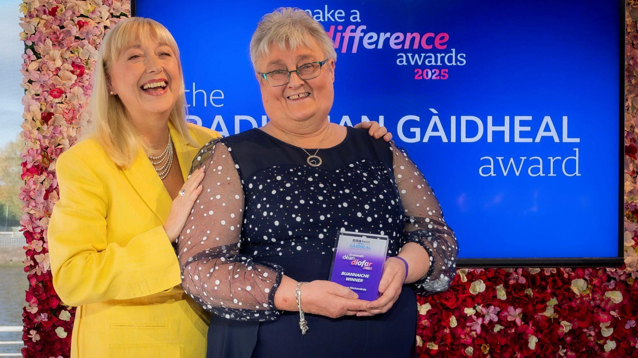 Seonag Anderson and Cathy Macdonald are smiling as they stand in front of a TV showing the name of the category Seonag won.