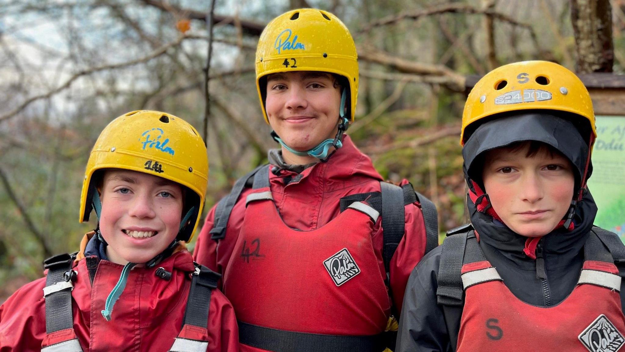 School friends Alfie, Ben and Jacob, smiling together at the camera. All are wearing red protective clothing and hard hats to go gorge walking.