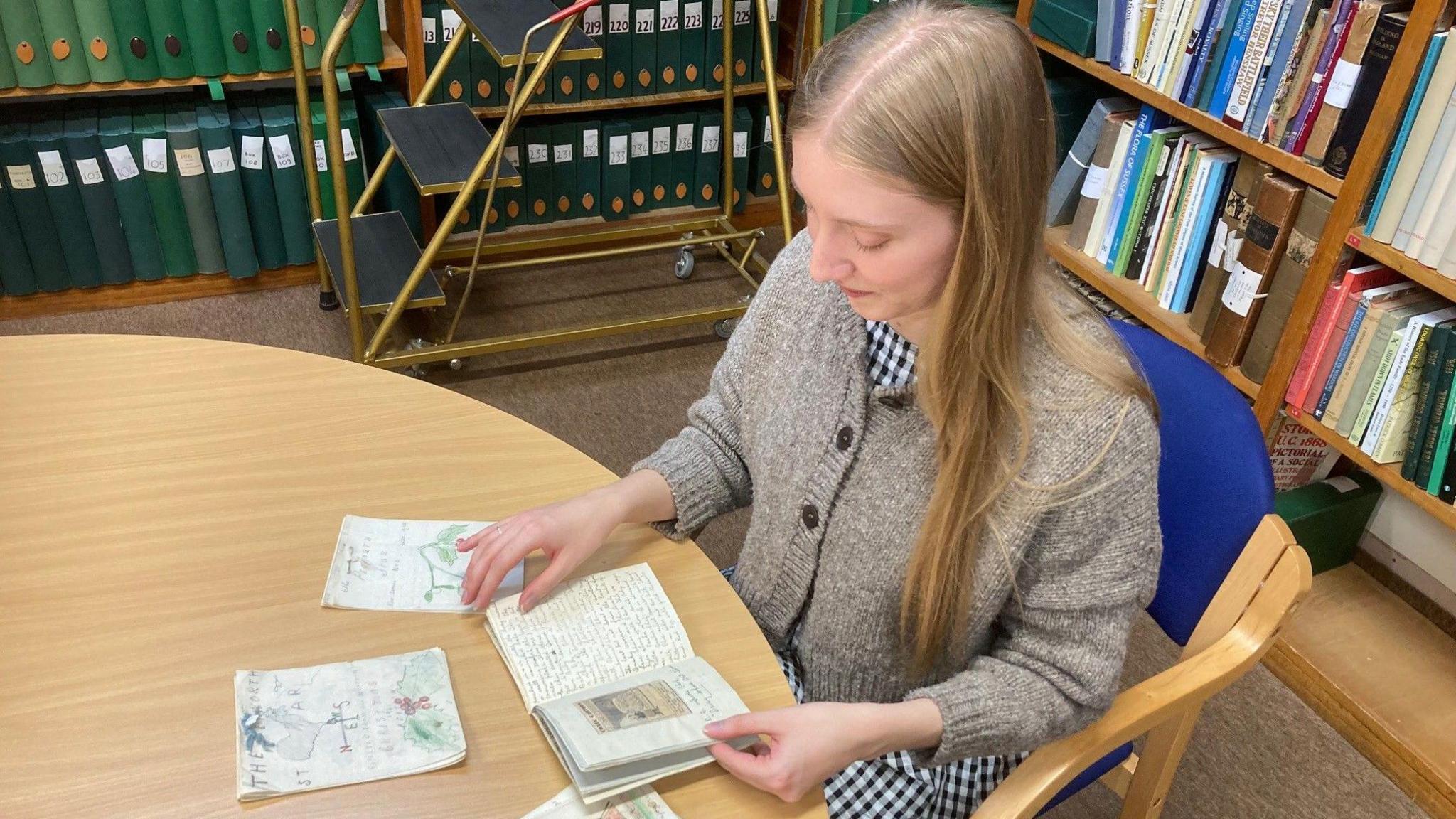 A photo taken above archivist Victoria Evans, dressed in a brown jumper and black and white gingham skirt, sat at a table, turning the pages of one of the tiny books lying in front of her on a wooden table. They are all hand-illustrated and handwritten in what looks like pencil. Behind her are bookshelves loaded with books and folders.