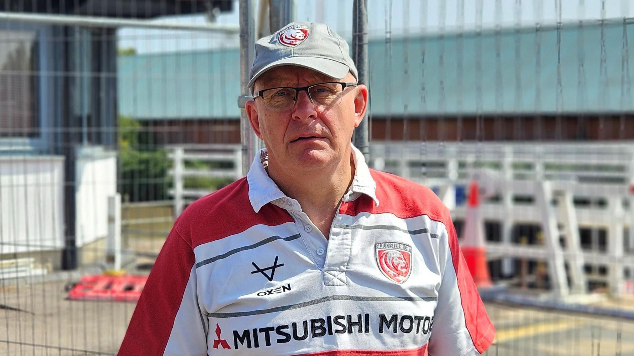 Dave Bucknell in a red and white Mitsubishi collared t-shirt. He is also wearing a grey cap with a red logo on it and is standing in front of a building site.