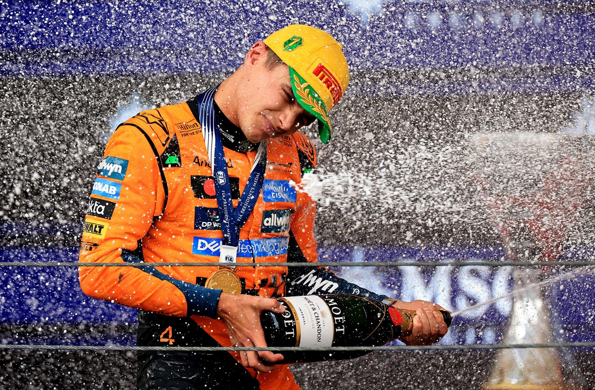 Person in an orange racing suit and cap spraying champagne from a large bottle during a podium celebration, with water droplets creating a dynamic splash effect against a backdrop of sponsor logos and a metallic trophy partially visible.