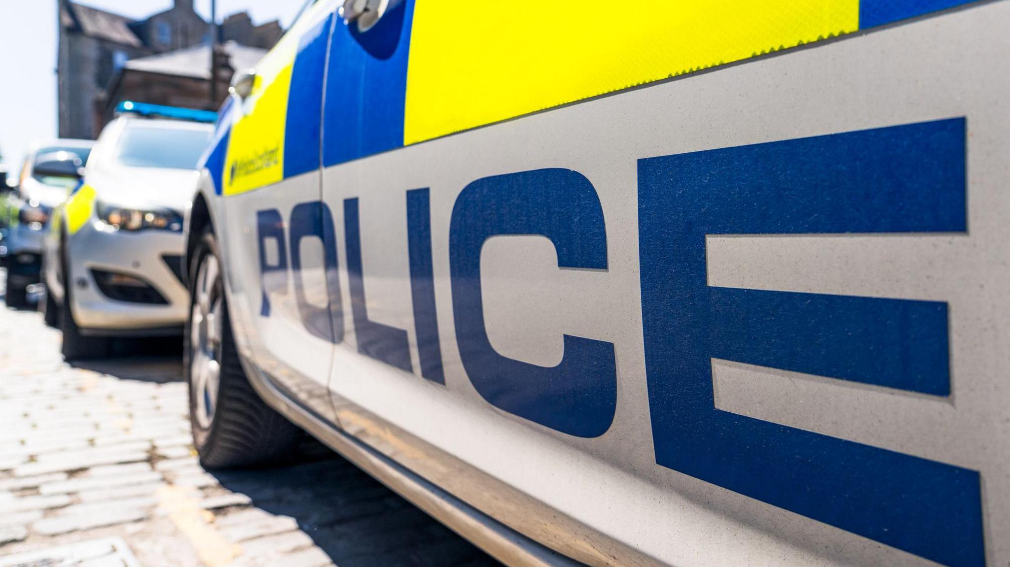 The word 'Police' below the fluorescent decals on a police car on a UK street, with other police vehicles in the distance.