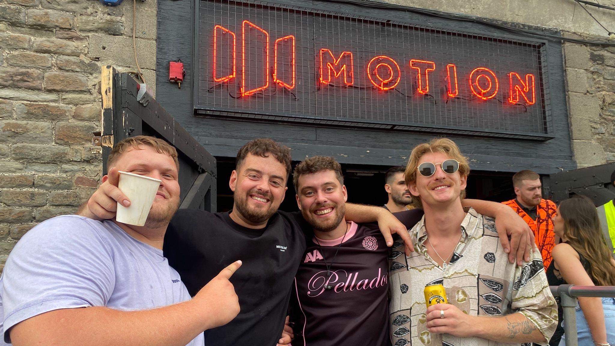 Four young men stand in the outside area at Motion nightclub in Bristol. They are smiling and it is a sunny afternoon. Above them the word "Motion" is spelt out in red illuminated letters. The wall of the building is made of dark grey brick.